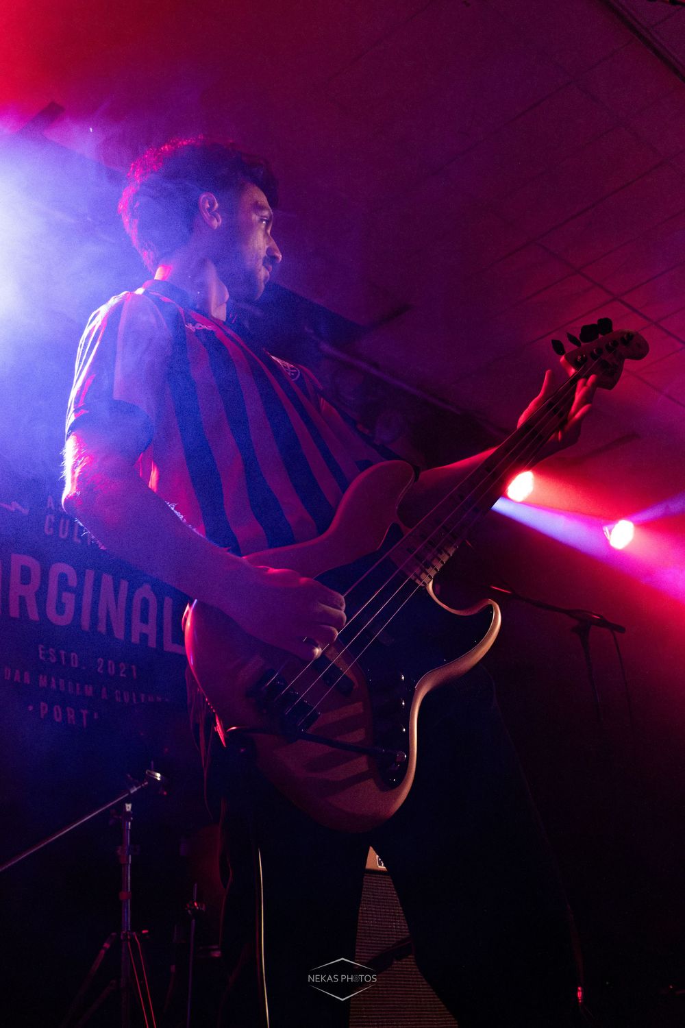 Bassist performing on stage under red and pink lights wearing a striped shirt at a live music concert.