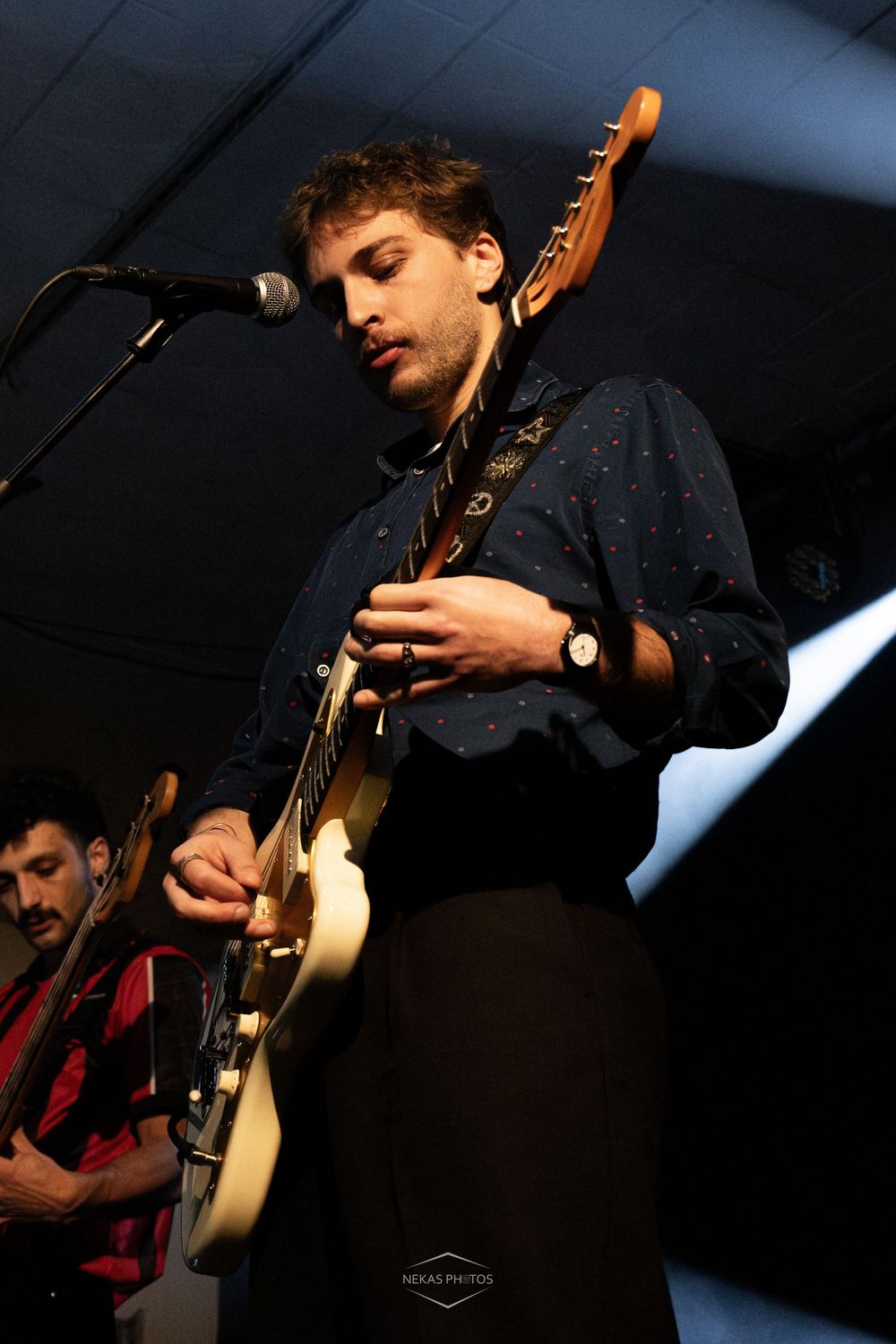 Young male guitarist performing on stage, playing a white electric guitar while standing at a microphone in a dark blue shirt.