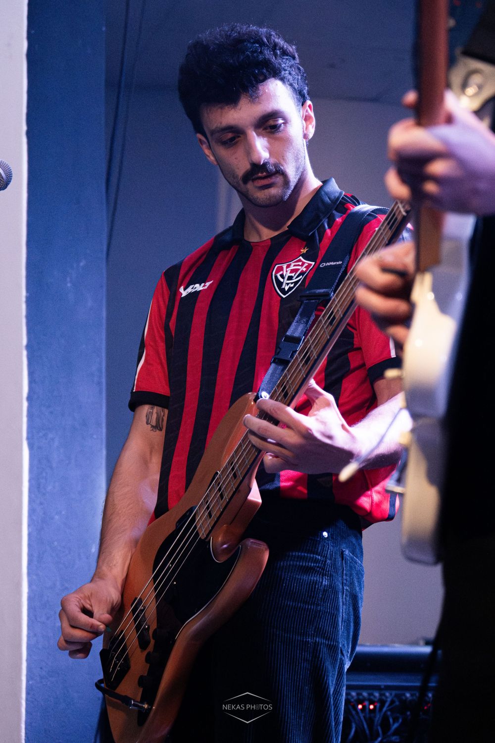 Young male bassist in red and black striped jersey performs on stage under blue and pink stage lighting.