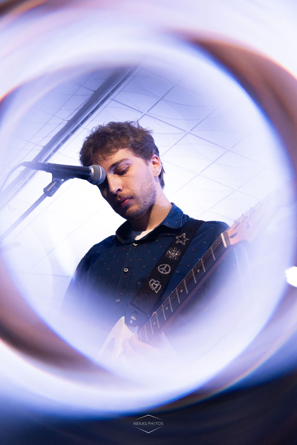 Young male guitarist performing on stage, captured through a circular lens flare effect with blue lighting.