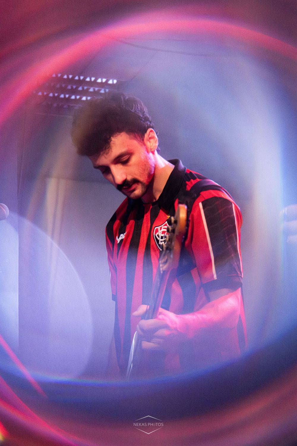 A young man with a mustache wearing a red and black striped soccer jersey, photographed through a circular bokeh frame.
