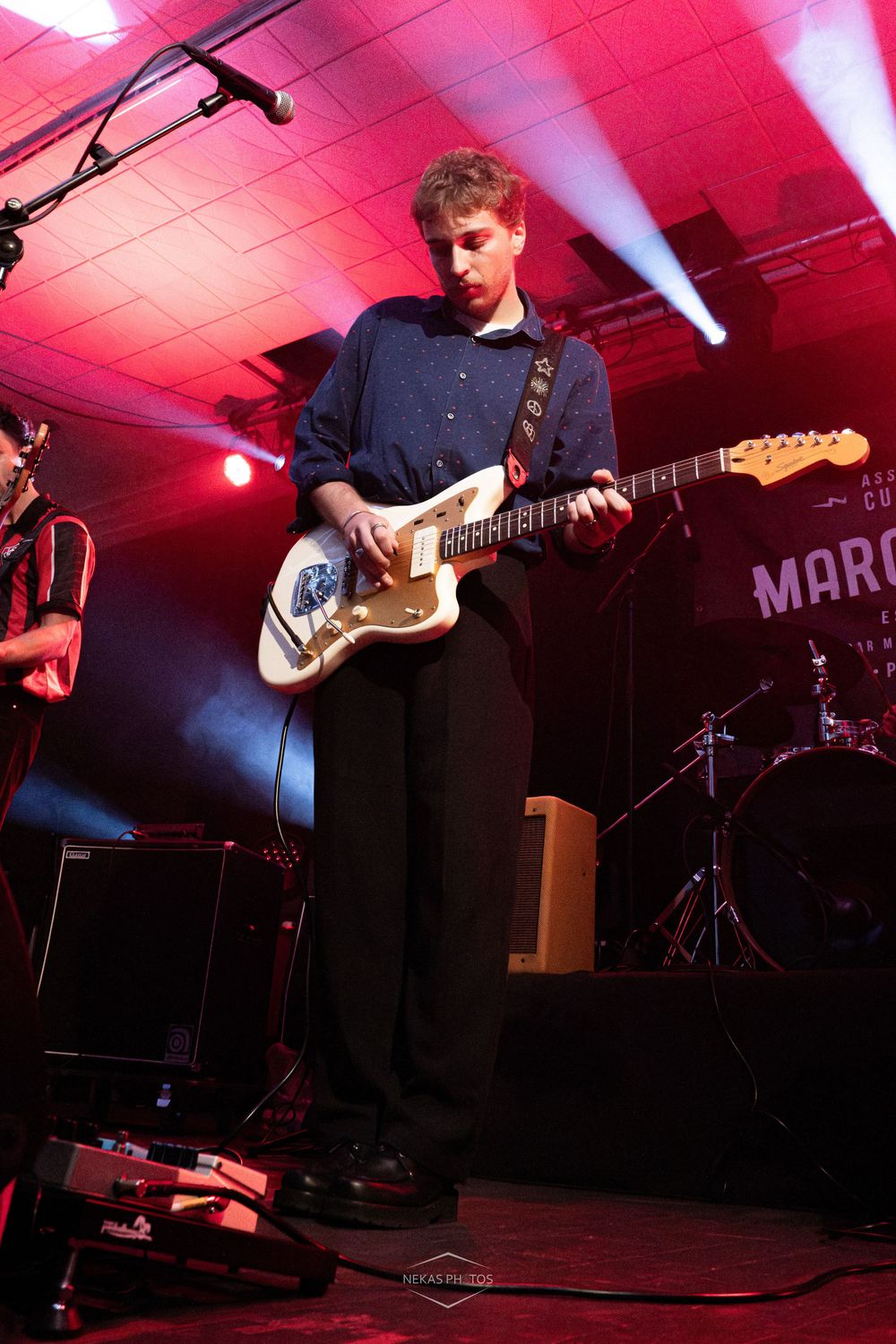 Young guitarist performing on stage with a white Fender guitar under red and blue lights at a live music venue.