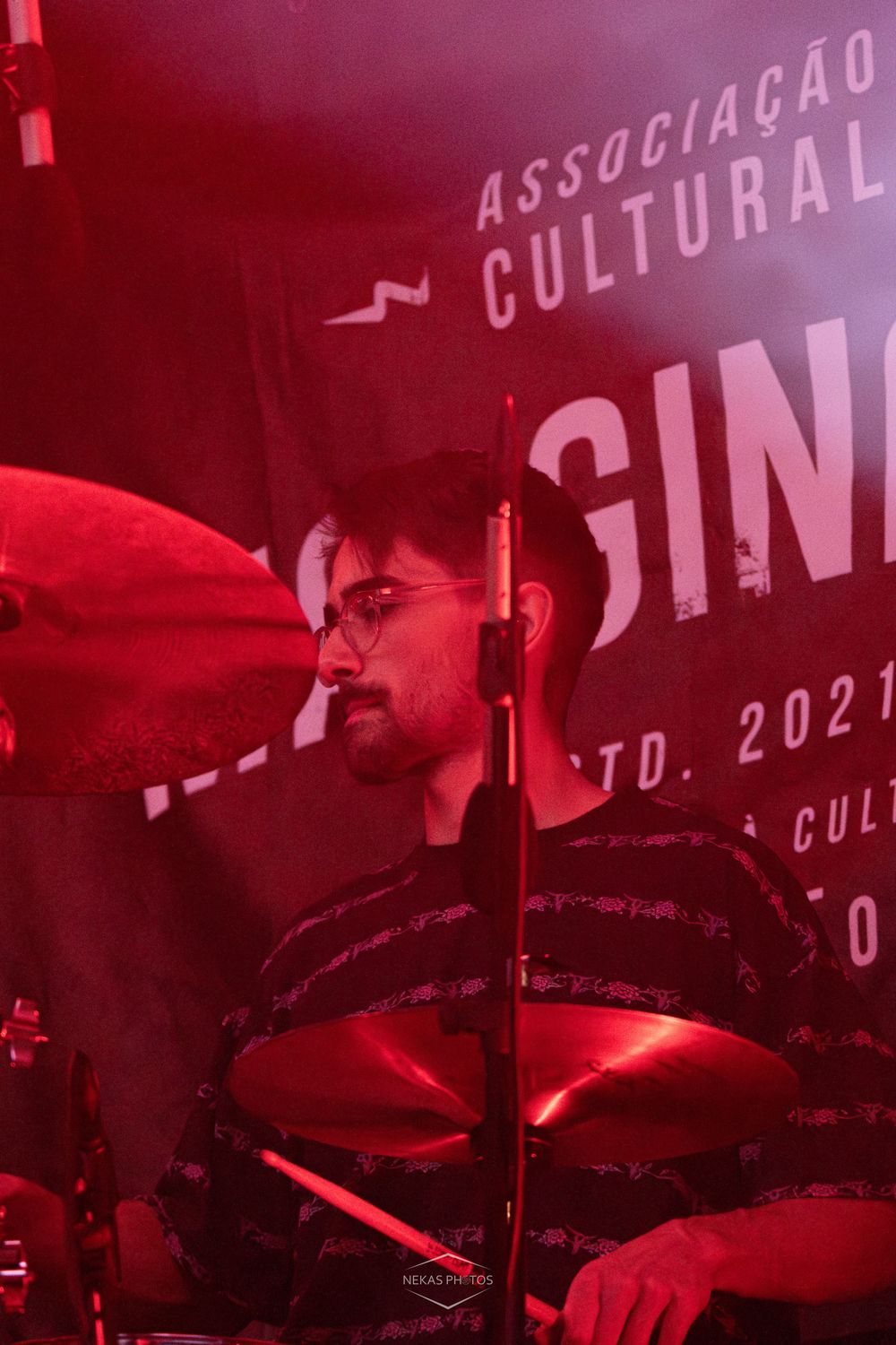 Male drummer performing live on stage under red lighting with cymbals and a cultural association banner behind him.