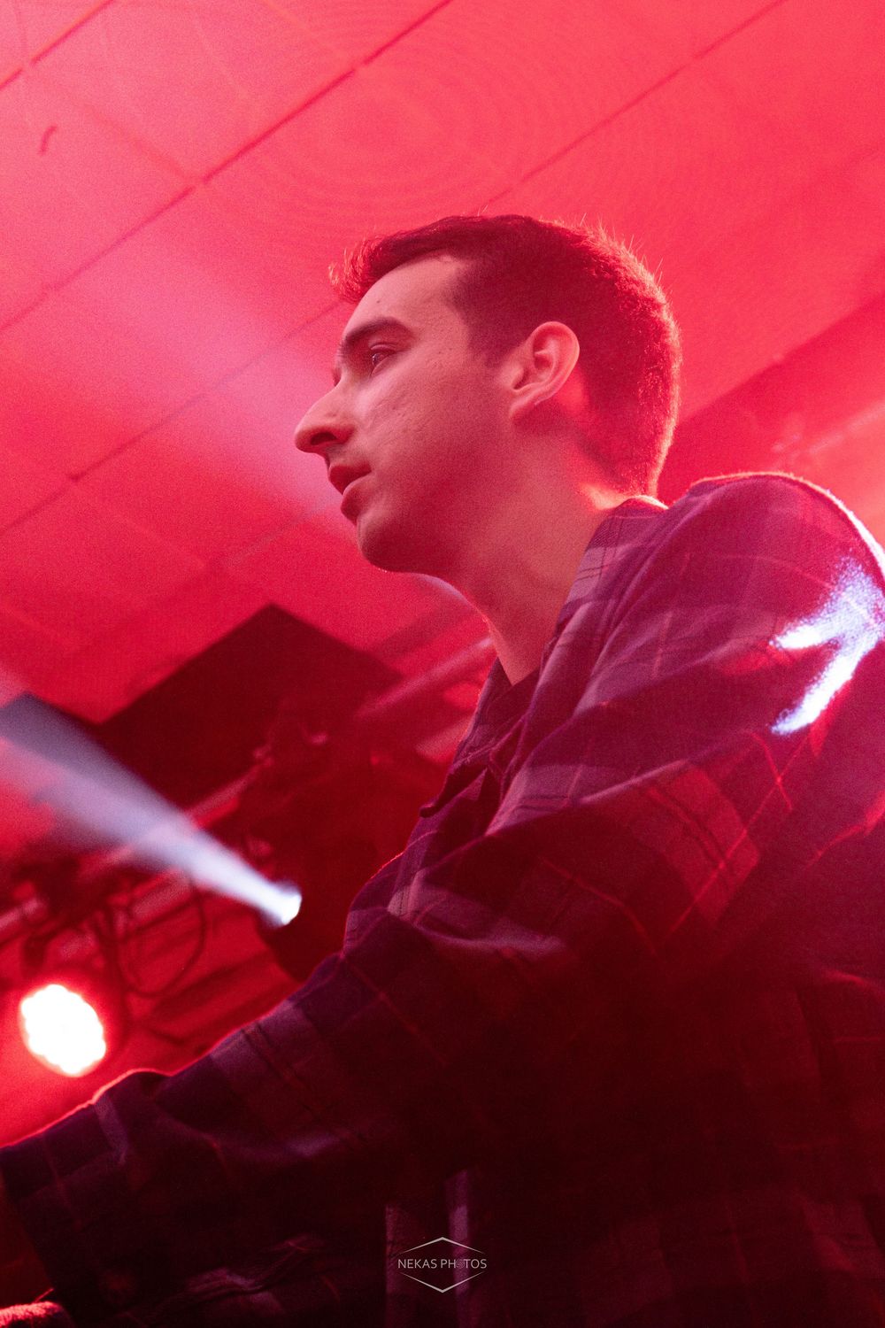 Young male DJ performing under red stage lights at a nightclub event, wearing a plaid shirt.