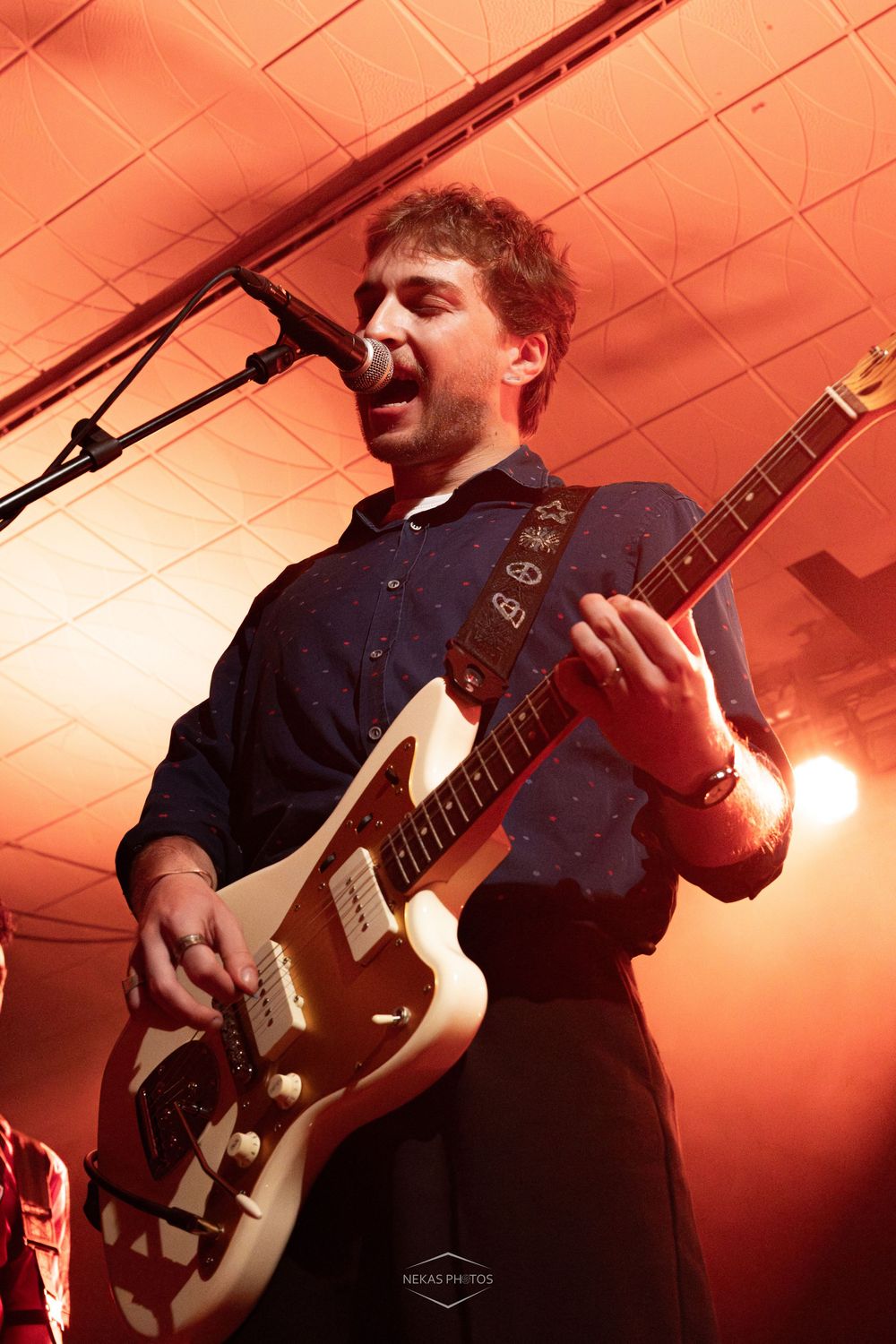 Young male guitarist performing on stage under red lights, singing into microphone while playing white electric guitar.