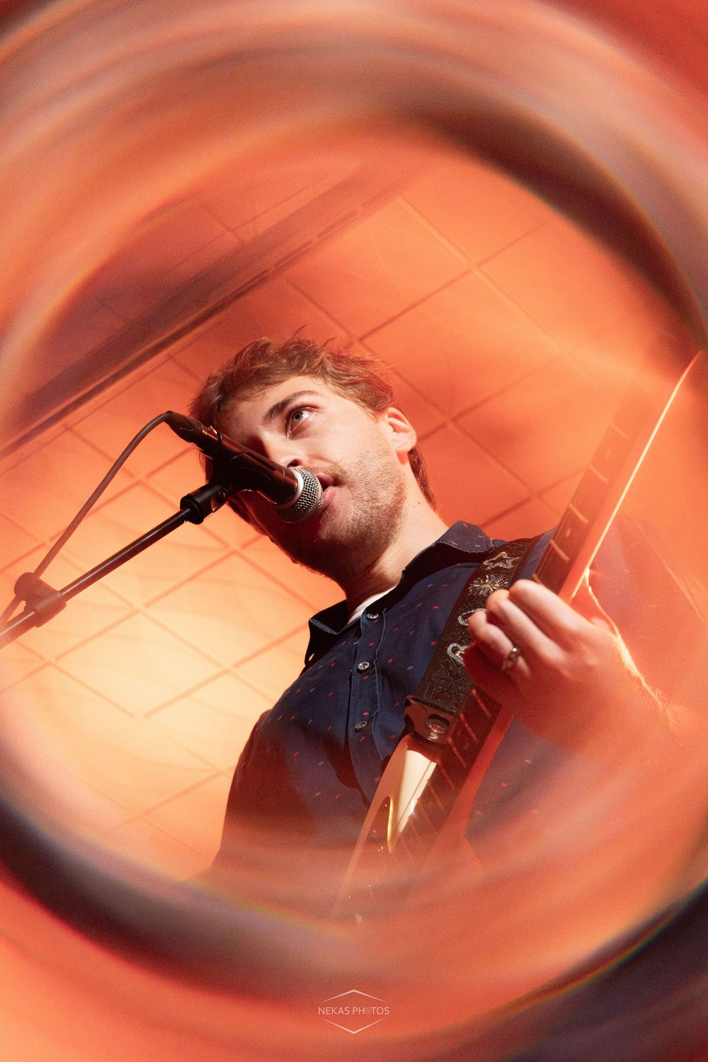 Young male musician singing into microphone while playing guitar, captured through creative swirling lens effect with warm red tones.
