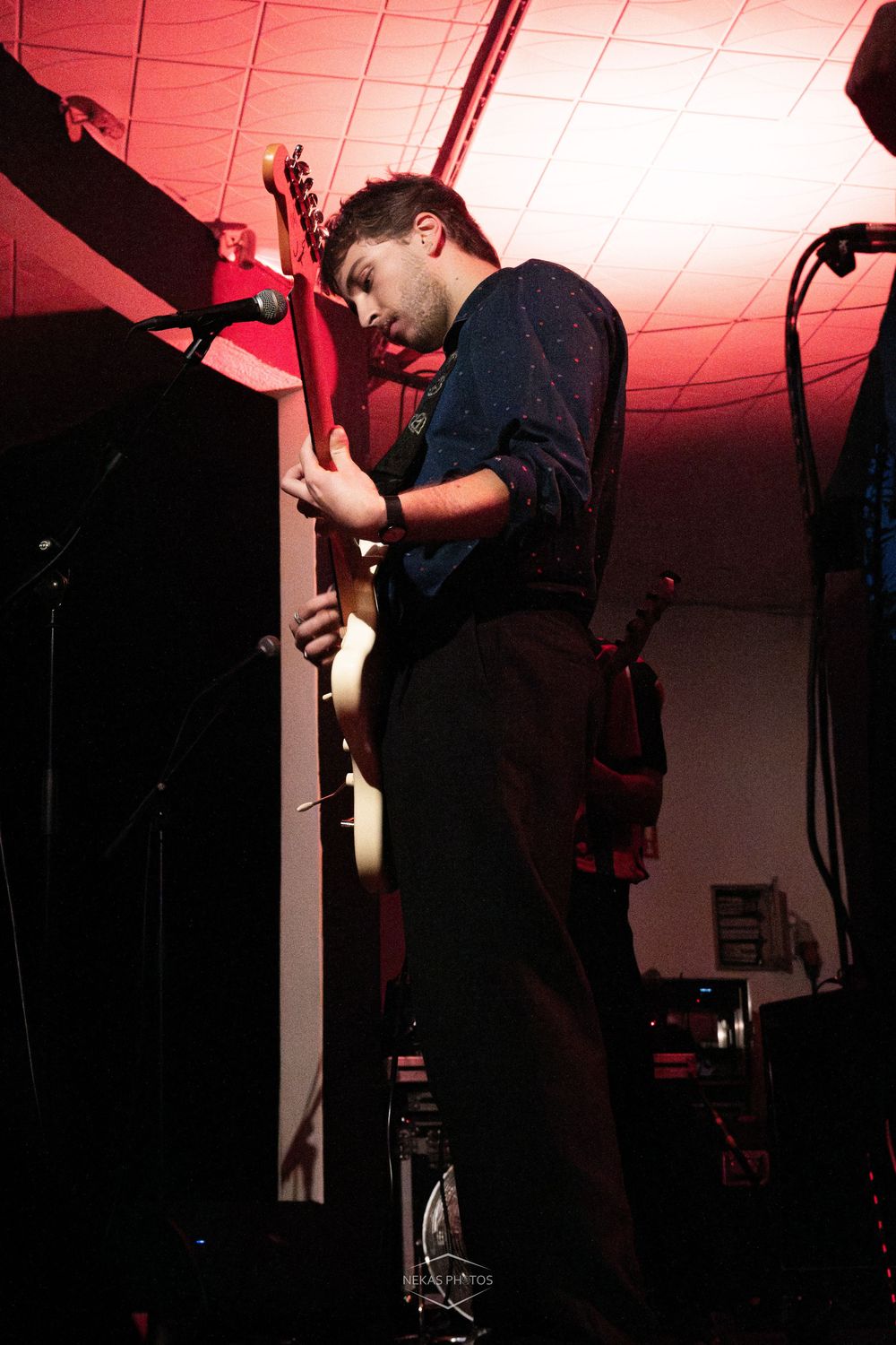 A young male guitarist performs on stage under red lighting, intensely focused while playing an electric guitar.
