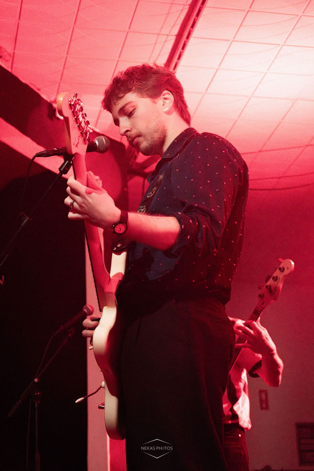 Young male guitarist performing on stage under red lighting at an indoor music venue.