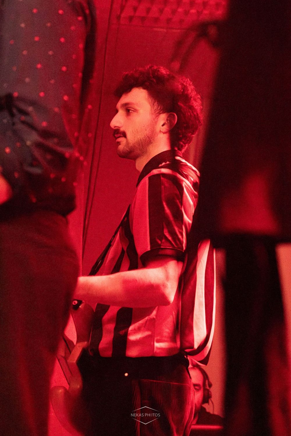 Man in striped shirt standing in red-lit venue, captured in profile view at a nightclub event.