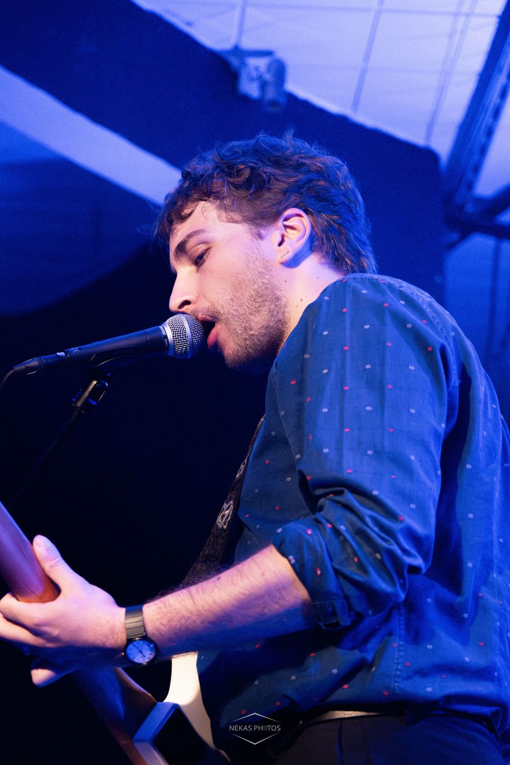Young male musician singing into microphone on stage, wearing dark blue dotted shirt and watch under blue stage lighting.
