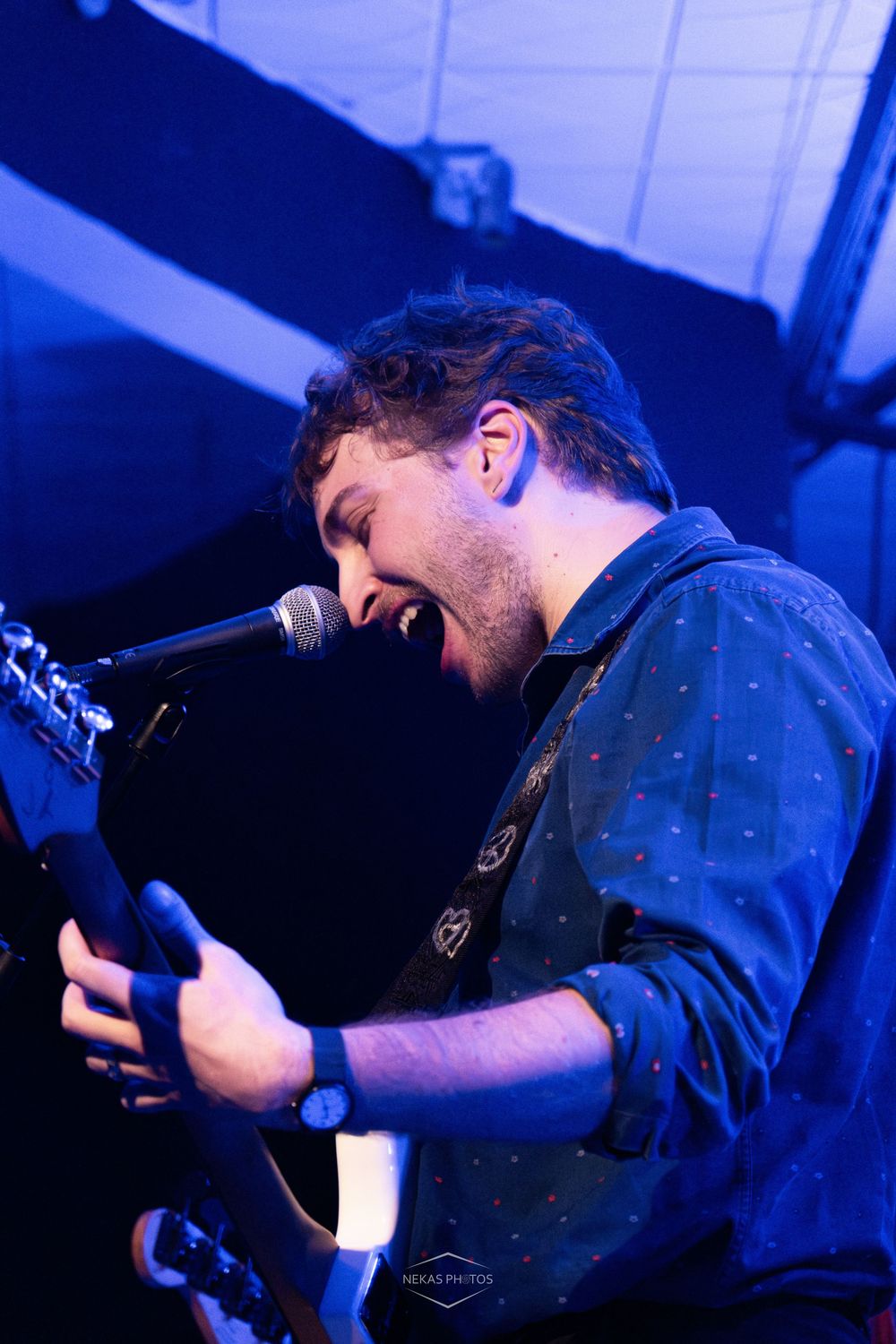 Young male guitarist singing passionately into microphone on stage under blue lights at live music concert.