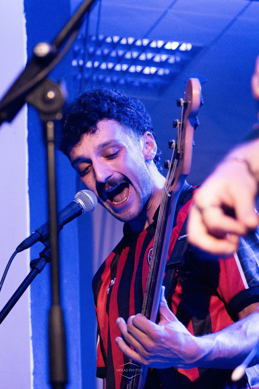 Musician singing and playing bass guitar on stage under blue lighting at a live music event.