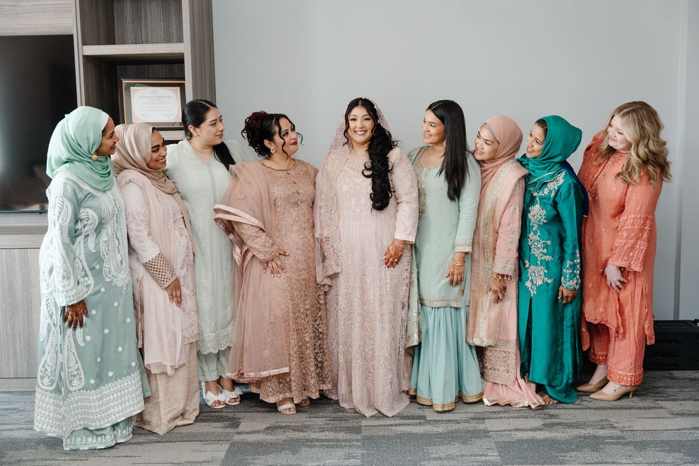 South Asian bride in blush lehenga posing with bridesmaids in colorful traditional outfits indoors.