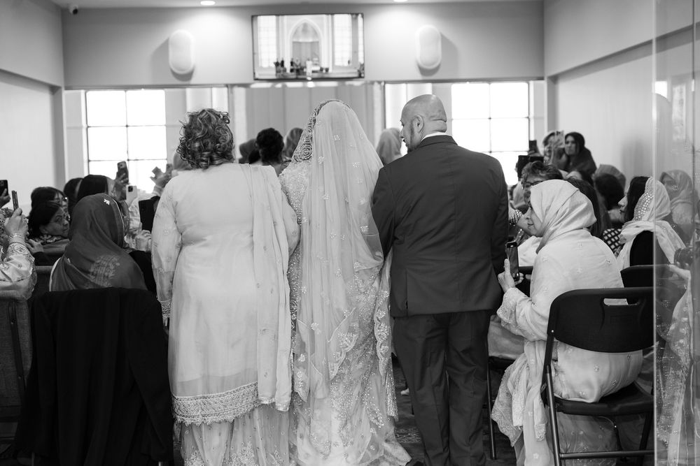 Black and white photo of South Asian bride in white gown walking down aisle with family at wedding.