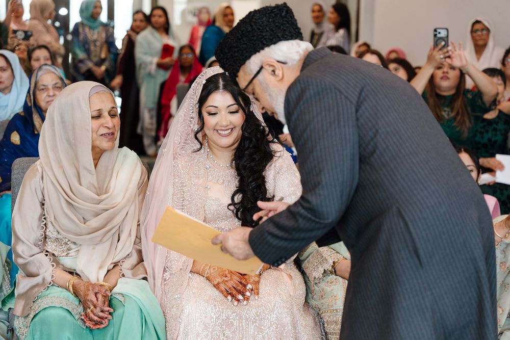 South Asian bride in blush lehenga smiling as elder relative blesses her during Nikah wedding ceremony.