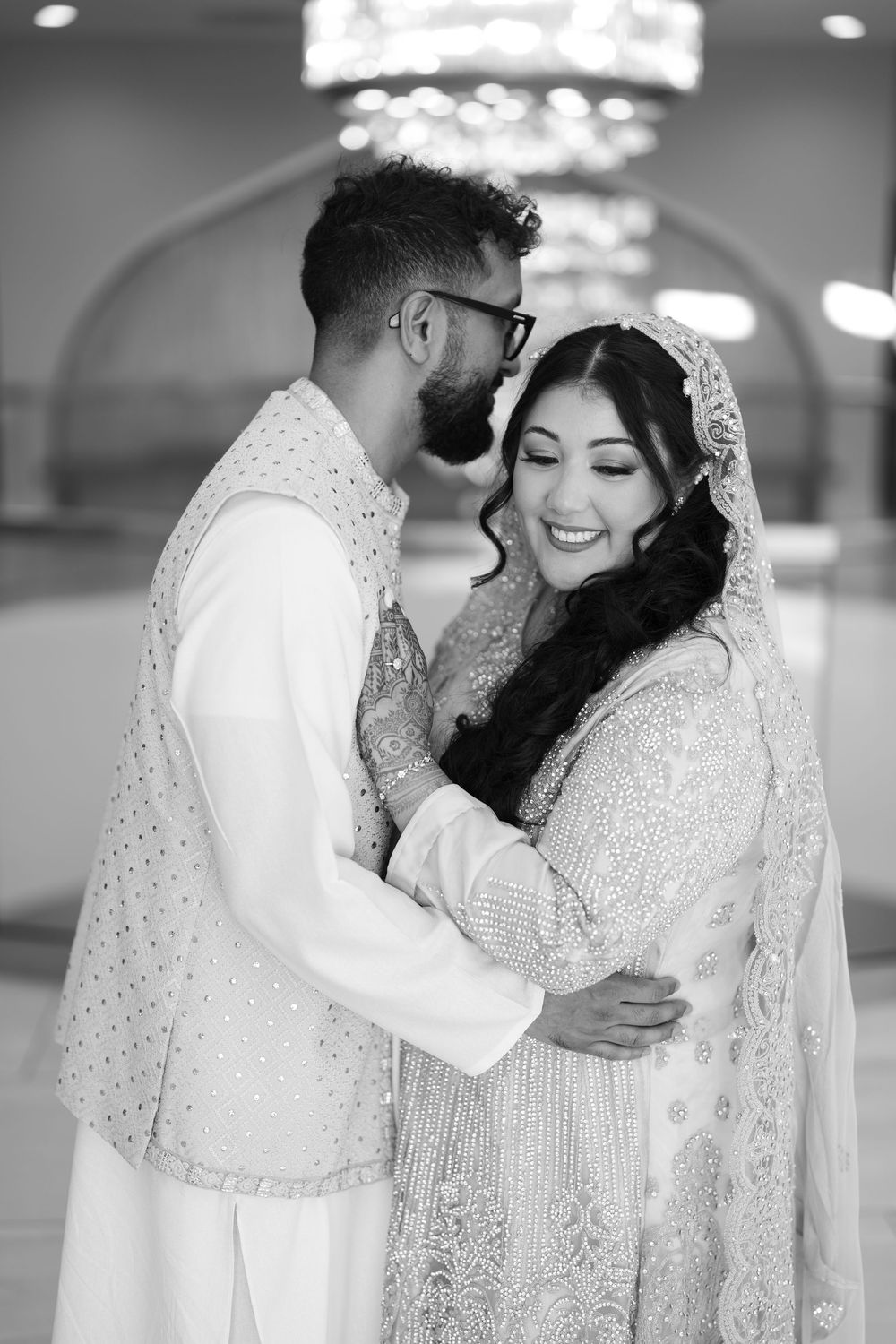 Black and white photo of South Asian bride and groom embracing and smiling in traditional wedding attire.