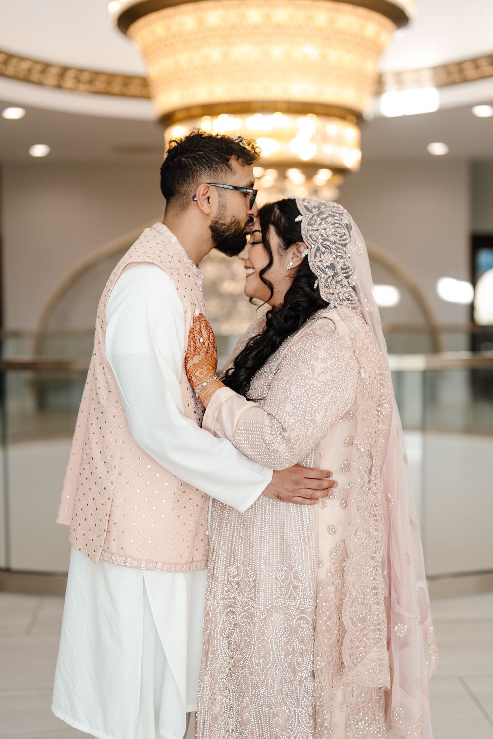 South Asian bride and groom in blush pink embroidered wedding attire embracing under grand chandelier.