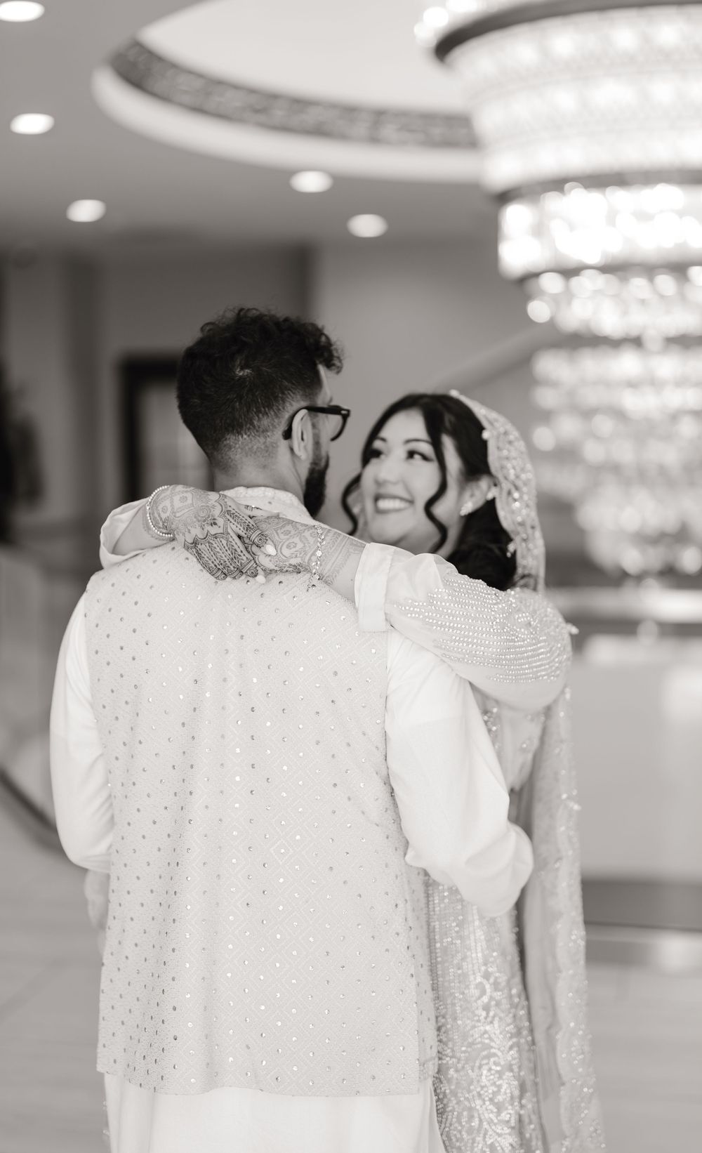 Black and white image of smiling South Asian bride embracing groom from behind under ornate chandelier.