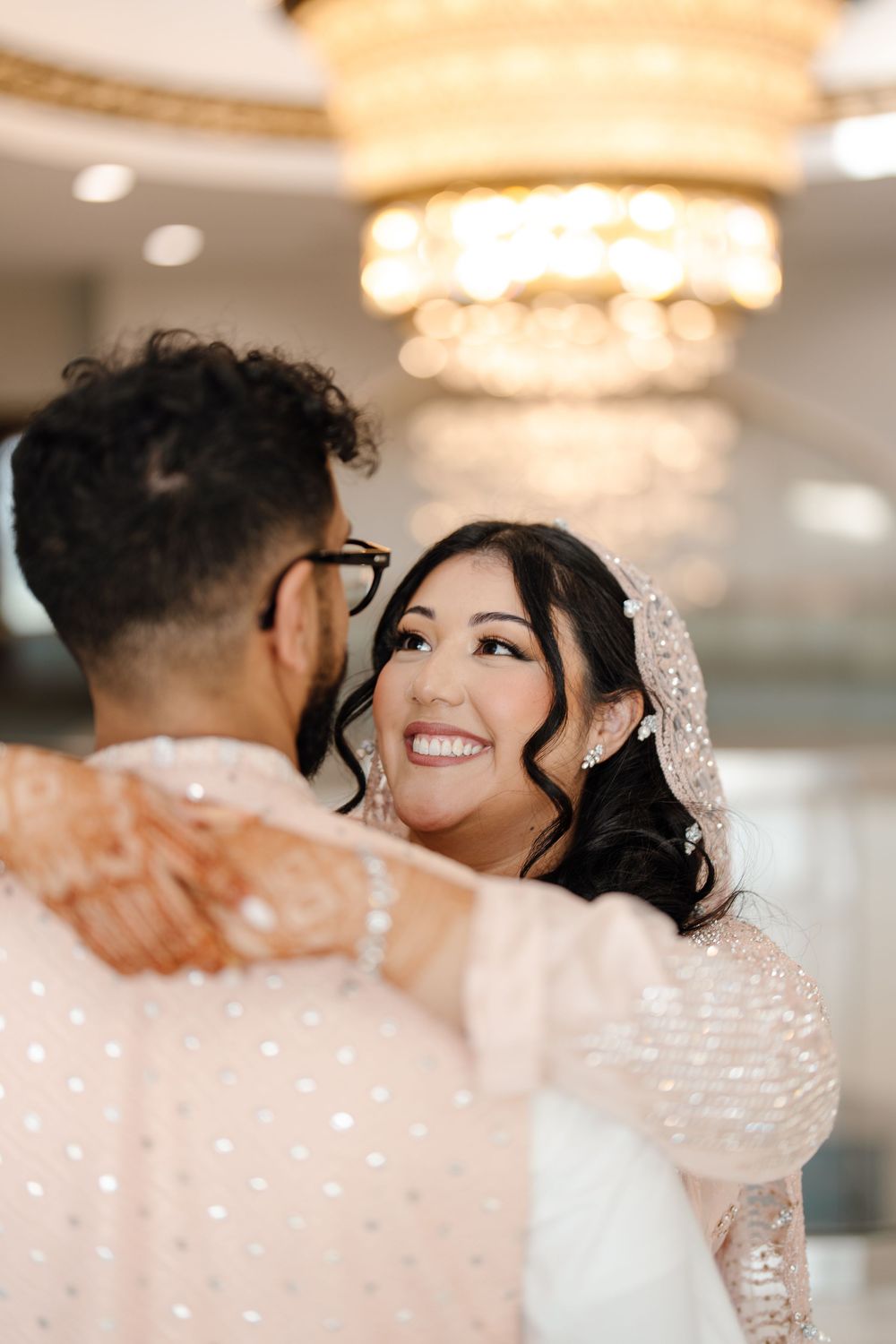 Smiling South Asian bride in blush embroidered lehenga and veil dancing with groom under chandelier.