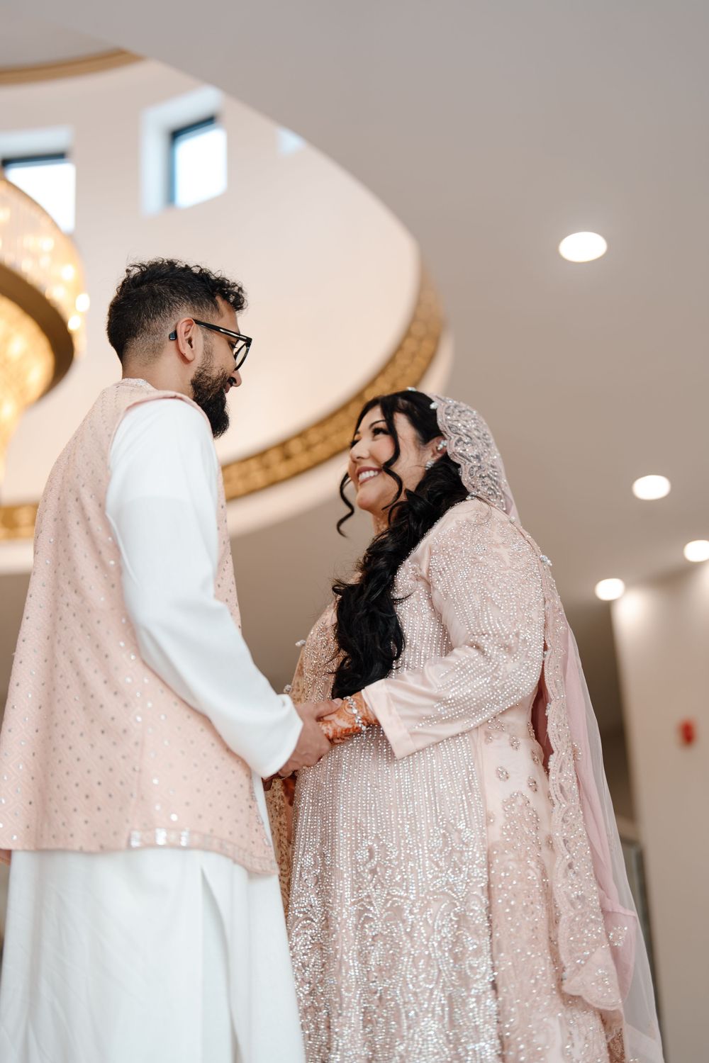 South Asian bride and groom in matching blush outfits holding hands and smiling at each other indoors.