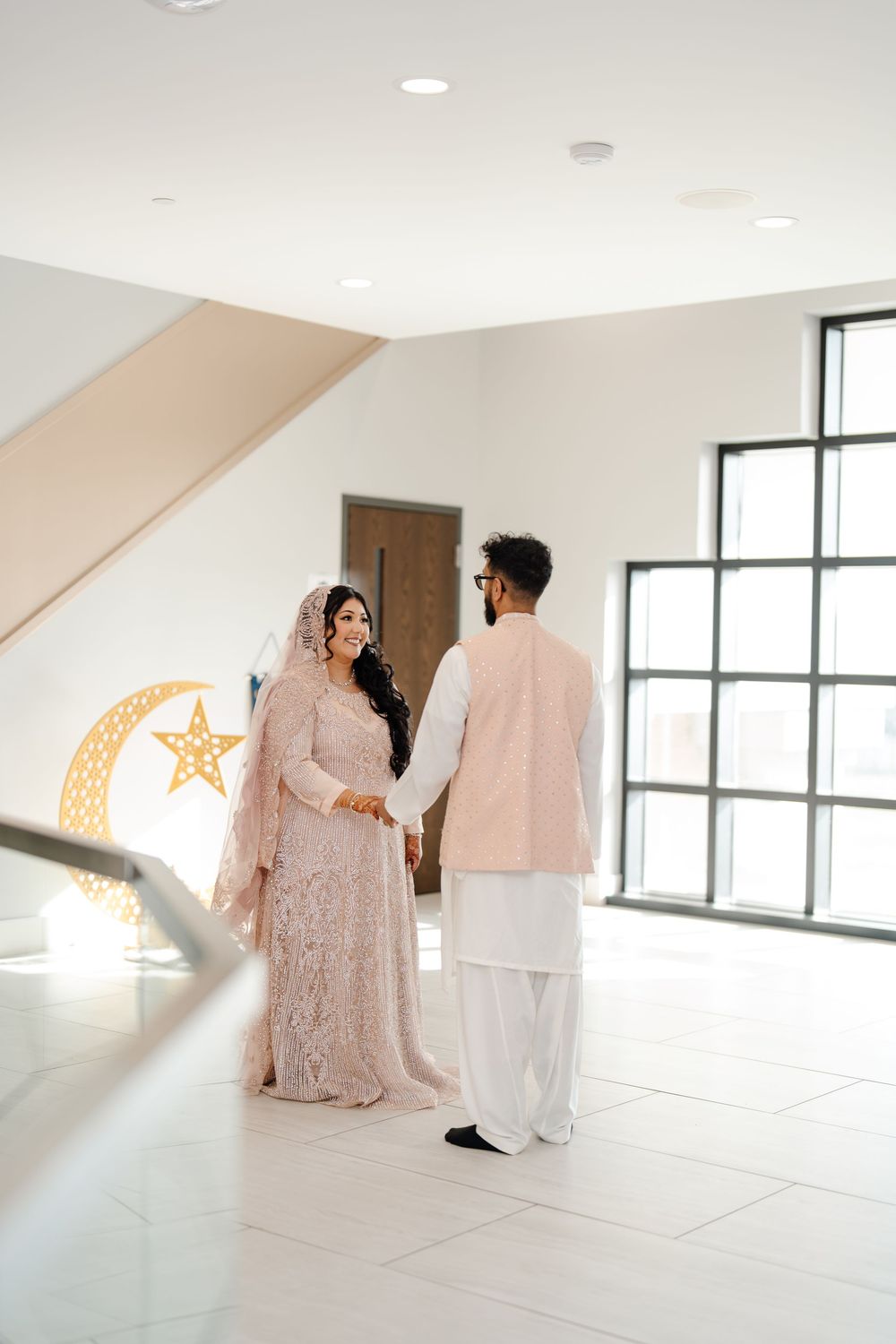 South Asian bride and groom in blush wedding attire standing together near crescent moon decor indoors.