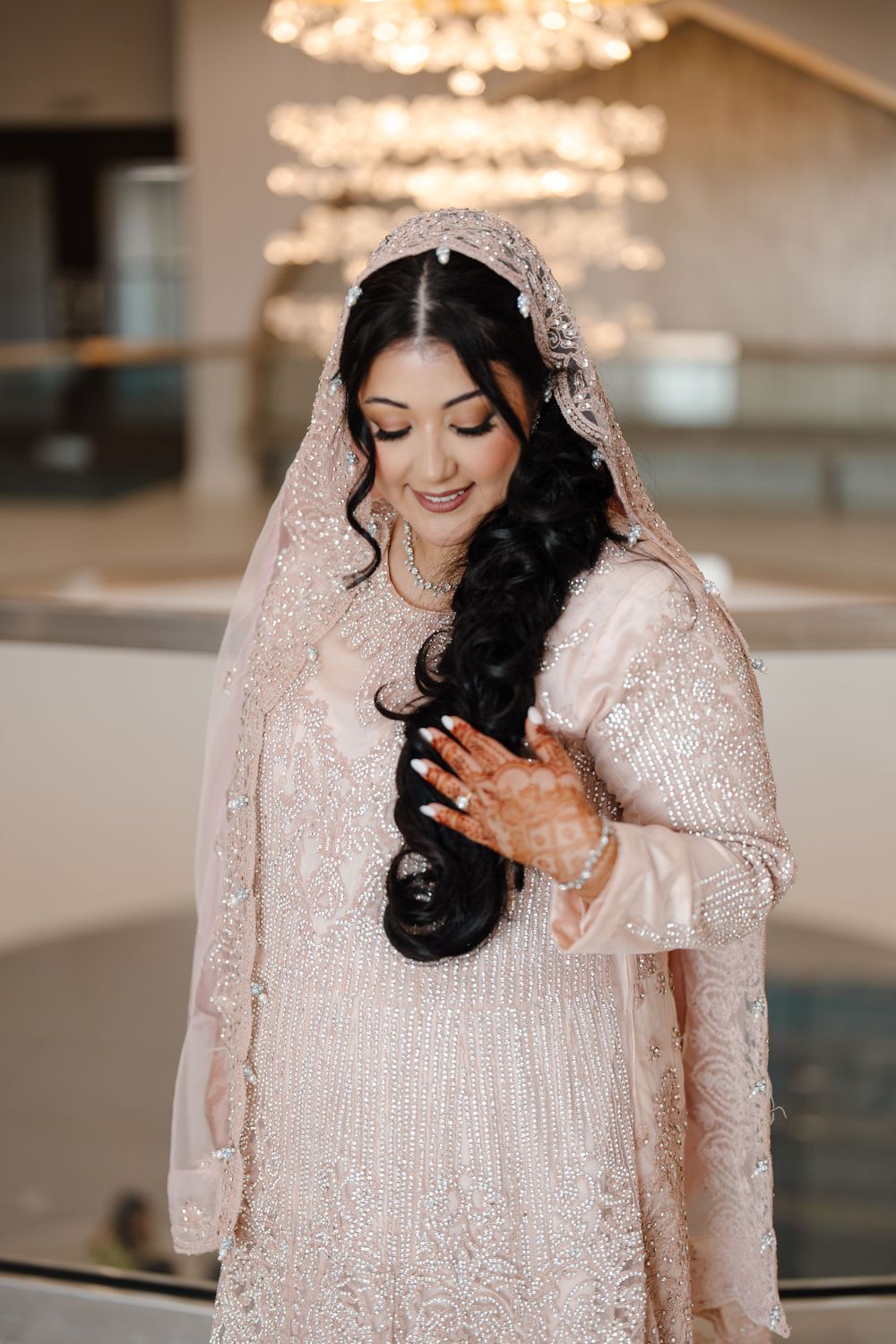 South Asian bride in blush embroidered lehenga and veil holding her braided hair under a chandelier.