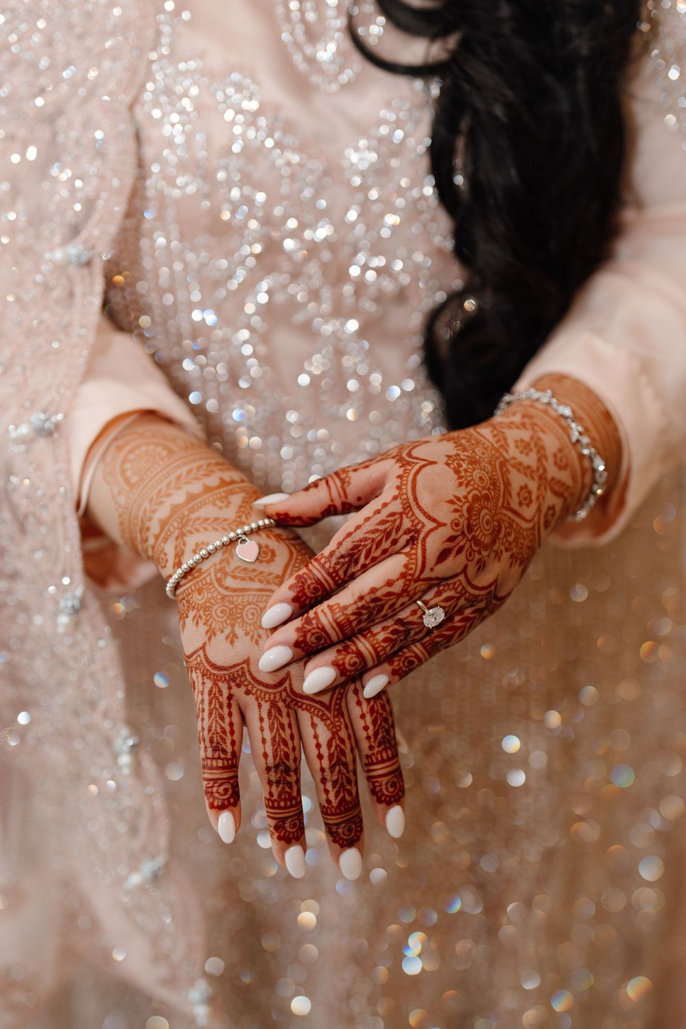 South Asian bride's hands displaying detailed red mehndi henna with bracelets on a sparkling lehenga.