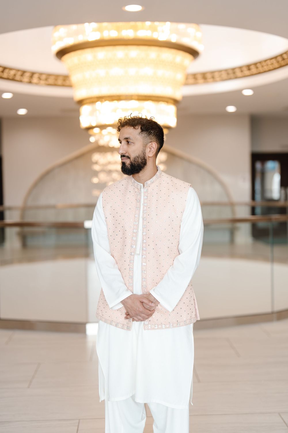 South Asian groom in white kurta and blush embroidered vest standing under a large gold chandelier.