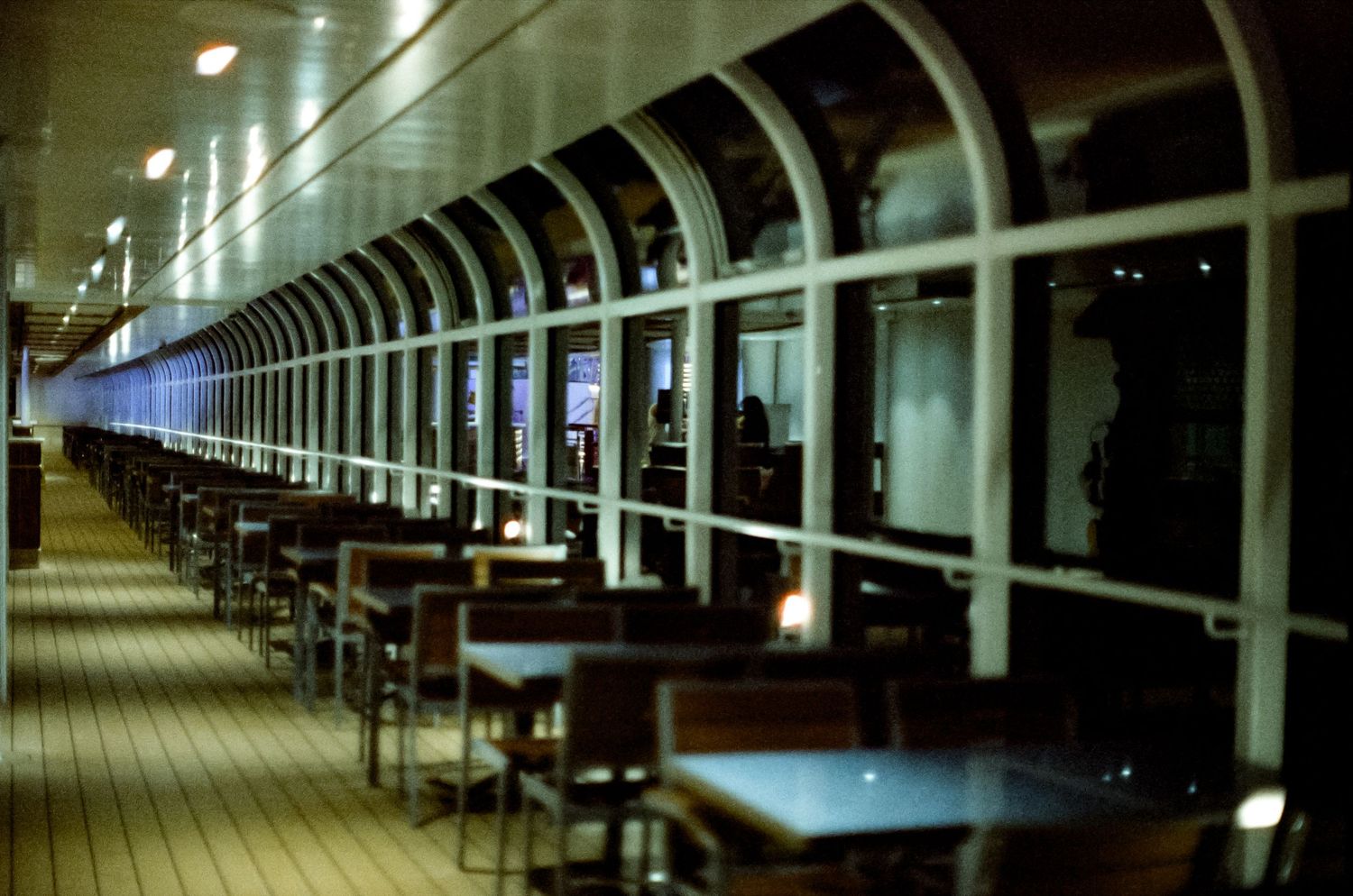 Empty outdoor deck dining area on a cruise ship at night, with arched windows and rows of wooden chairs and tables.