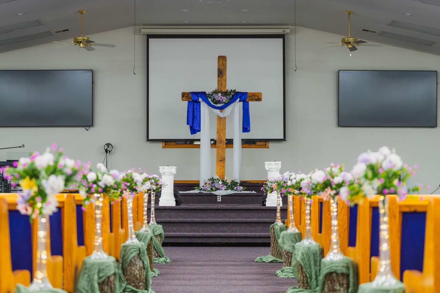 Church sanctuary decorated for a wedding with wooden cross, blue ribbon, white candles, floral arrangements, and yellow-trimmed pews.