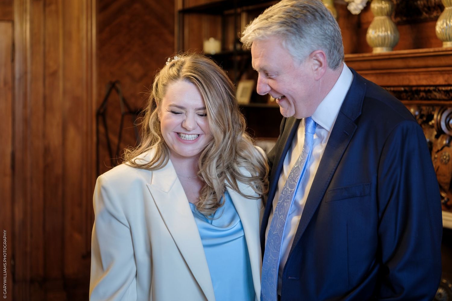 A woman in a beige blazer and man in a suit share a laugh together in an elegant wood-paneled room.