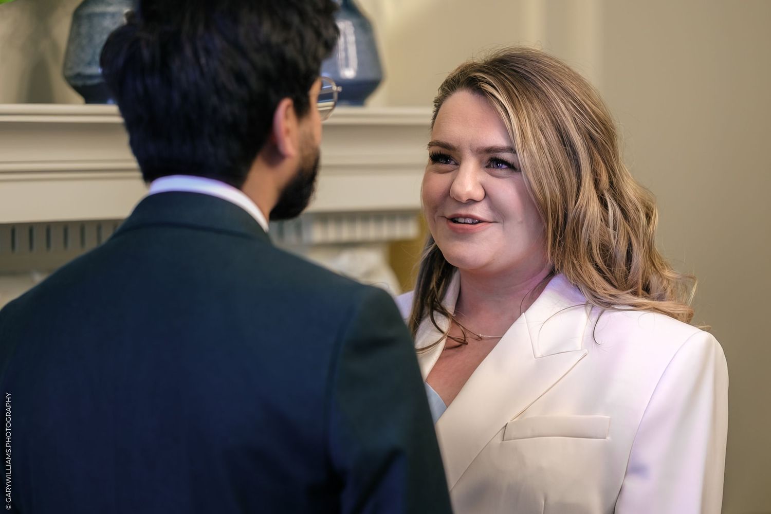A woman in a white blazer smiles while conversing with a man in a dark suit at a formal event.
