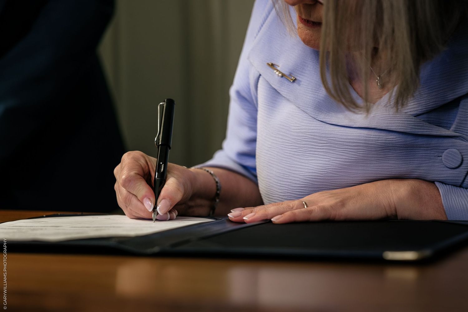 Woman in blue blazer signing official document with black pen at wooden desk.