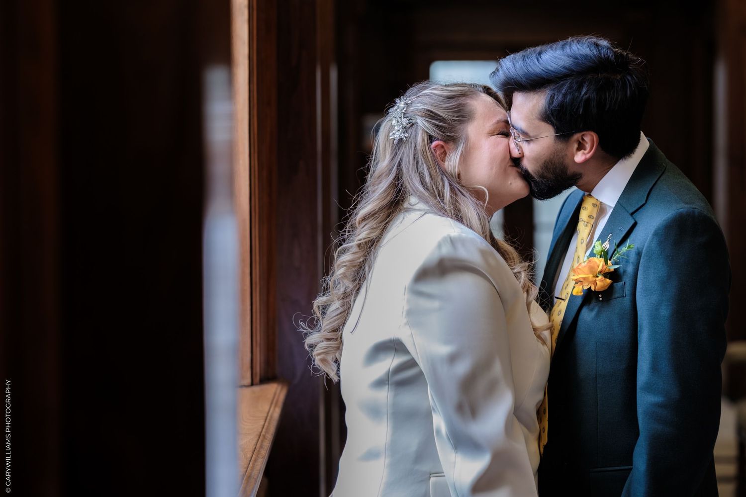 Bride and groom sharing a romantic kiss in a warmly lit hallway, bride in white jacket, groom in teal suit with yellow boutonniere.