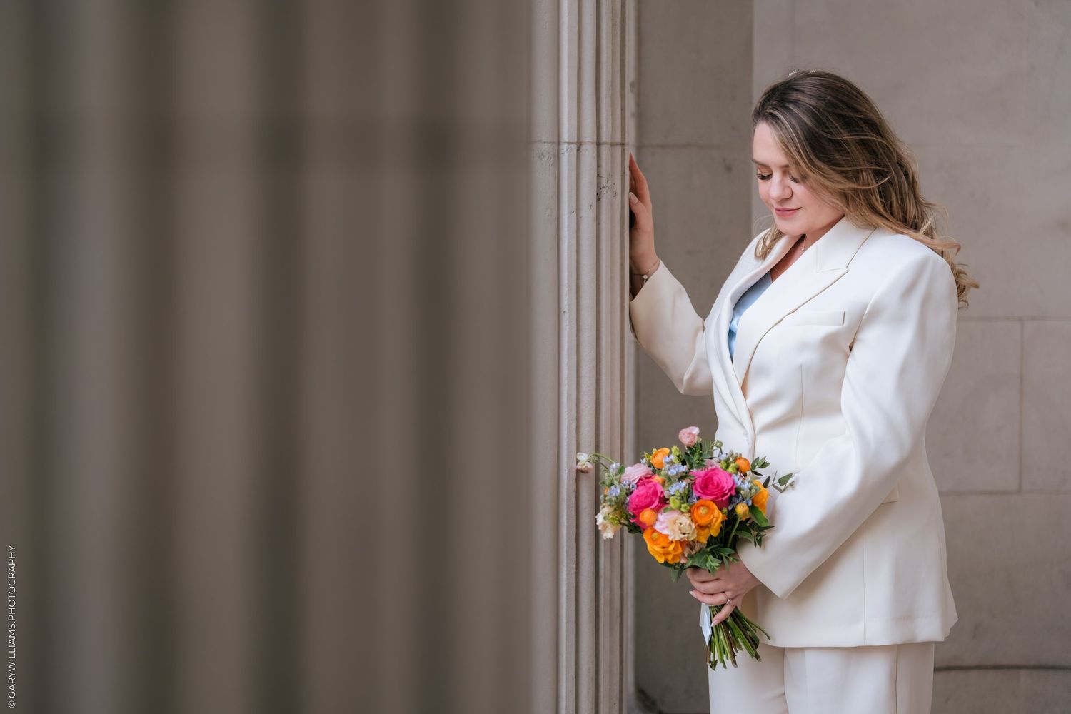 Bride in white suit holding colorful bouquet, leaning against stone column in elegant wedding portrait.
