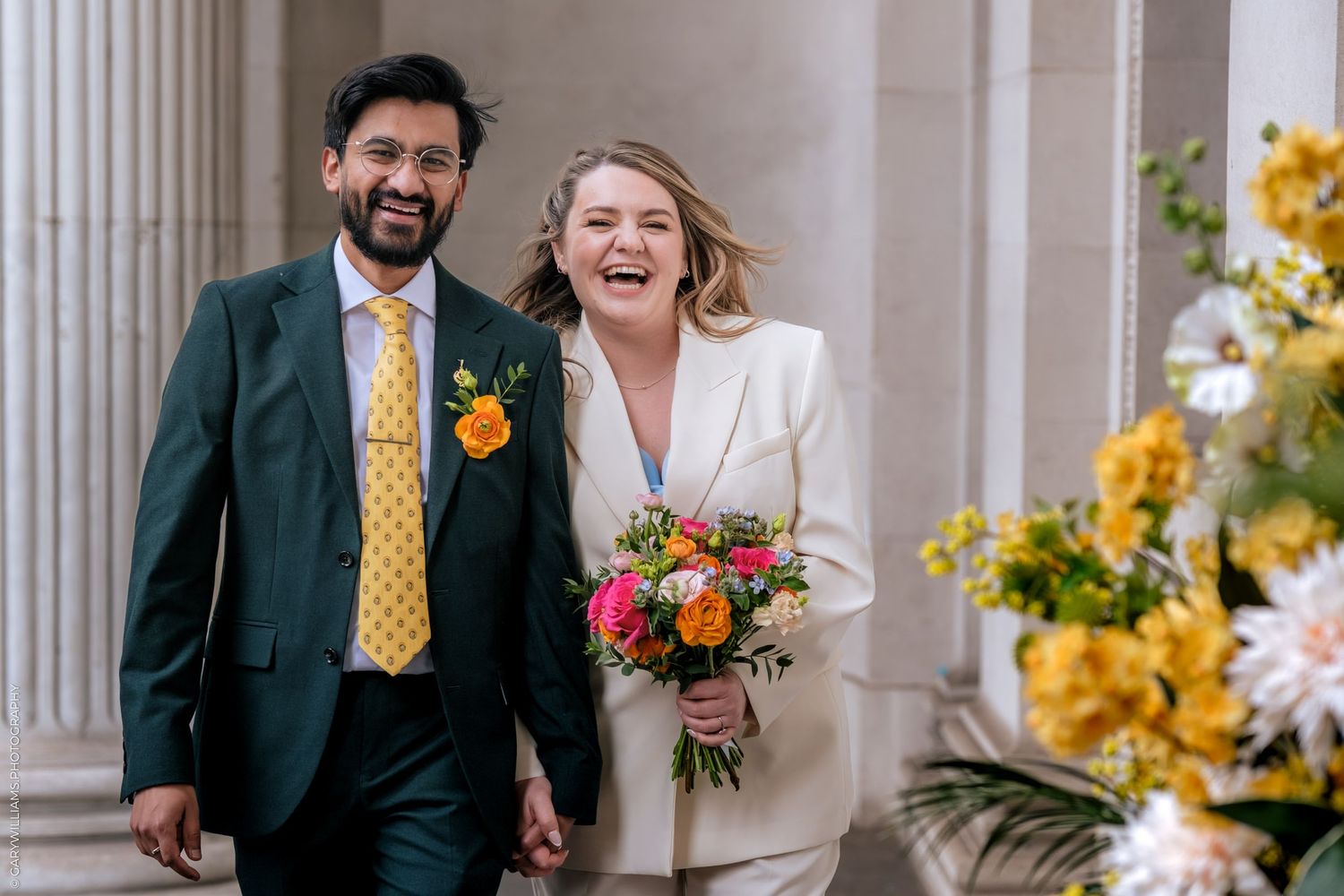 Happy couple laughing outside wedding venue, bride in white suit with colorful bouquet, groom in green suit with yellow tie.
