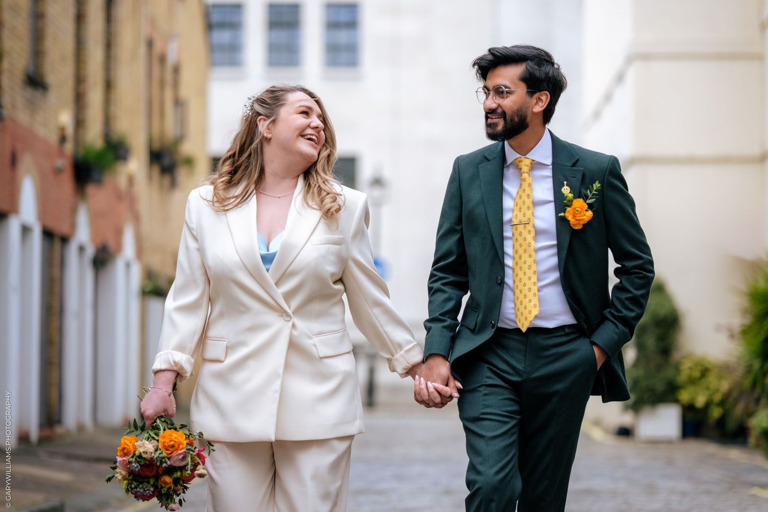 Happy couple in wedding attire holding hands and walking outdoors, bride in white suit holding bouquet, groom in green suit.