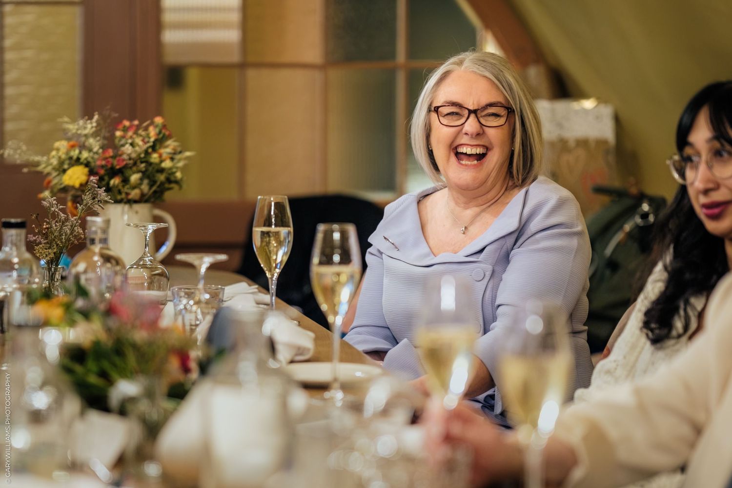 Woman laughing joyfully at an elegant dinner table with champagne glasses and floral centerpiece.