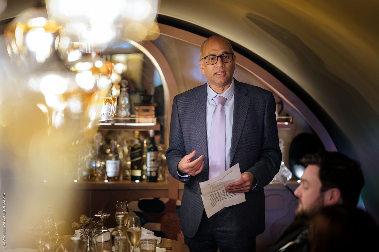 A man in a suit speaks at an upscale restaurant bar, holding papers, with warm lighting and bottles in background.