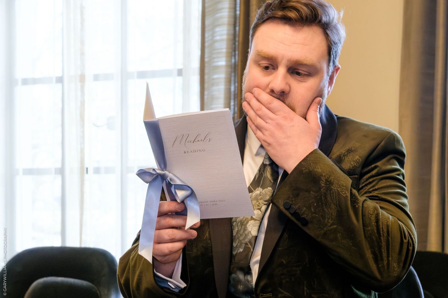 Man in velvet blazer reading a card with a bow, covering his mouth in surprise at a formal event.