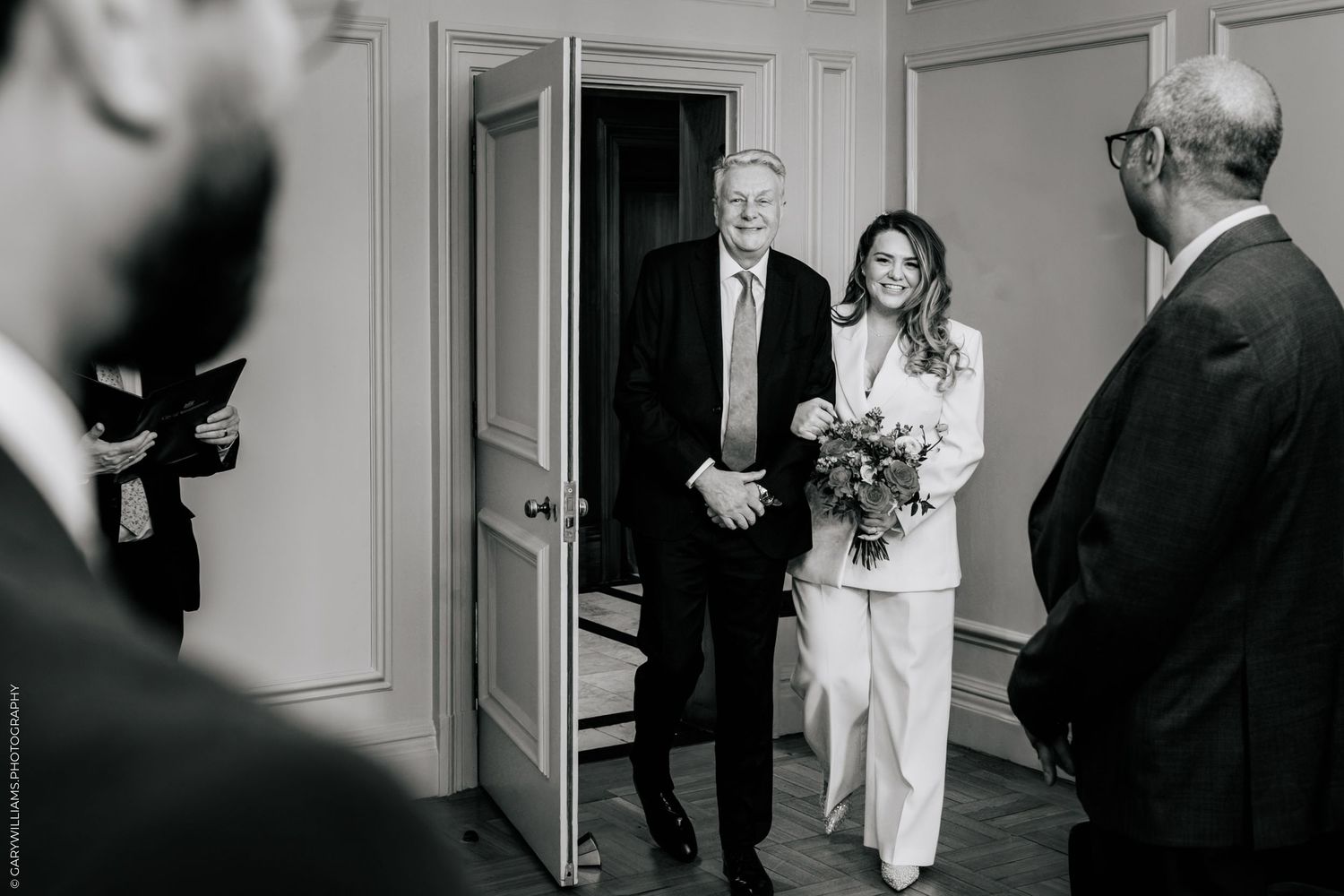 Black and white wedding photo of bride in white suit holding bouquet entering room with older man in suit.