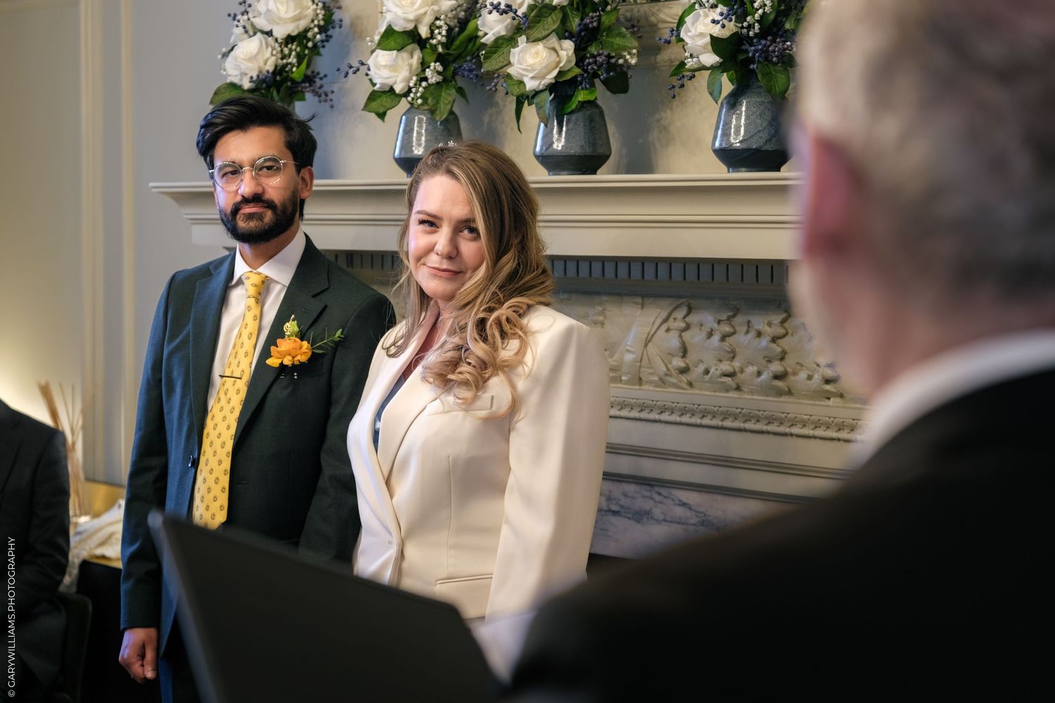 Bride and groom smiling during wedding ceremony, bride in white jacket, groom in grey suit with yellow tie.
