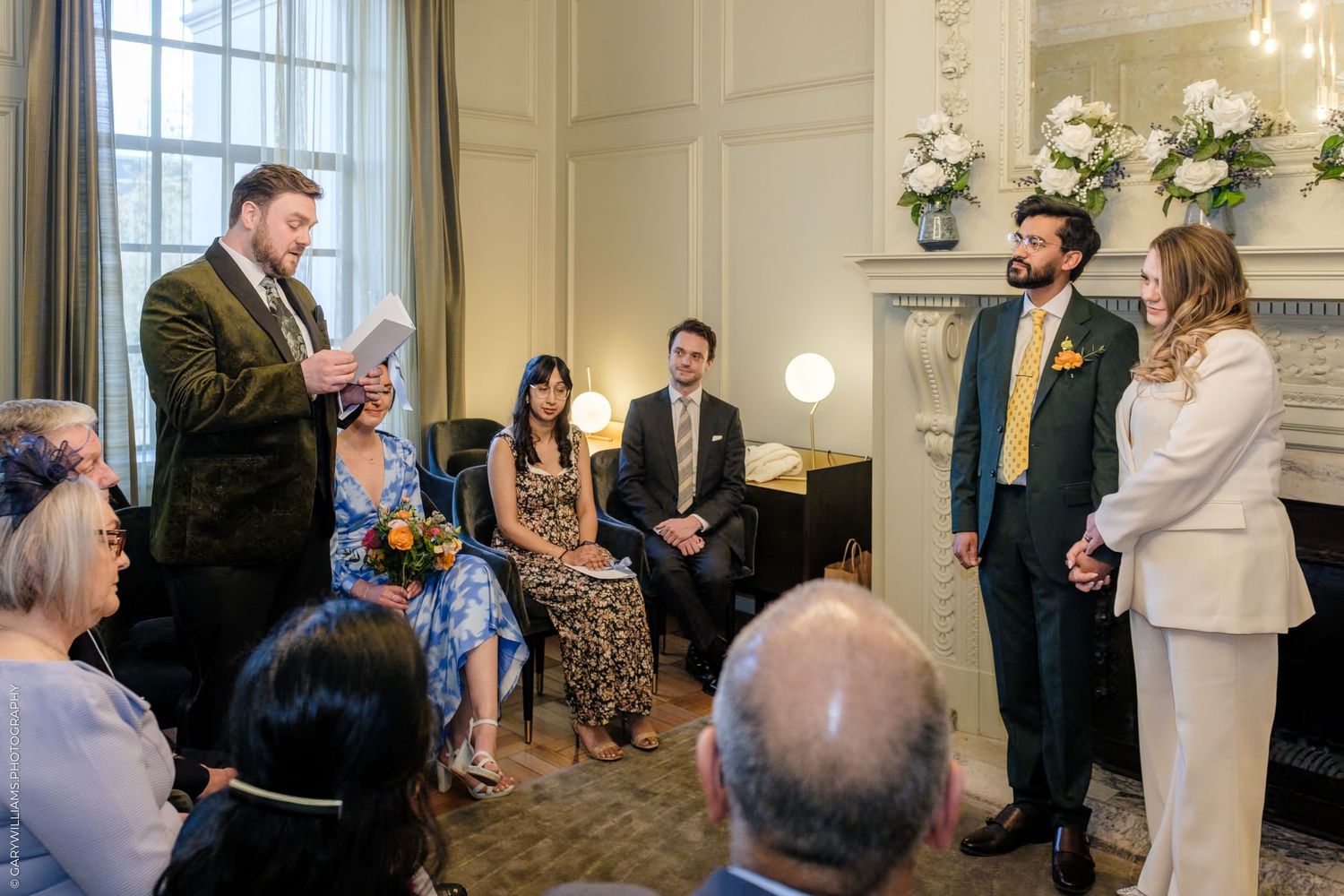 Intimate wedding ceremony in elegant room with couple exchanging vows before officiant and small group of seated guests.