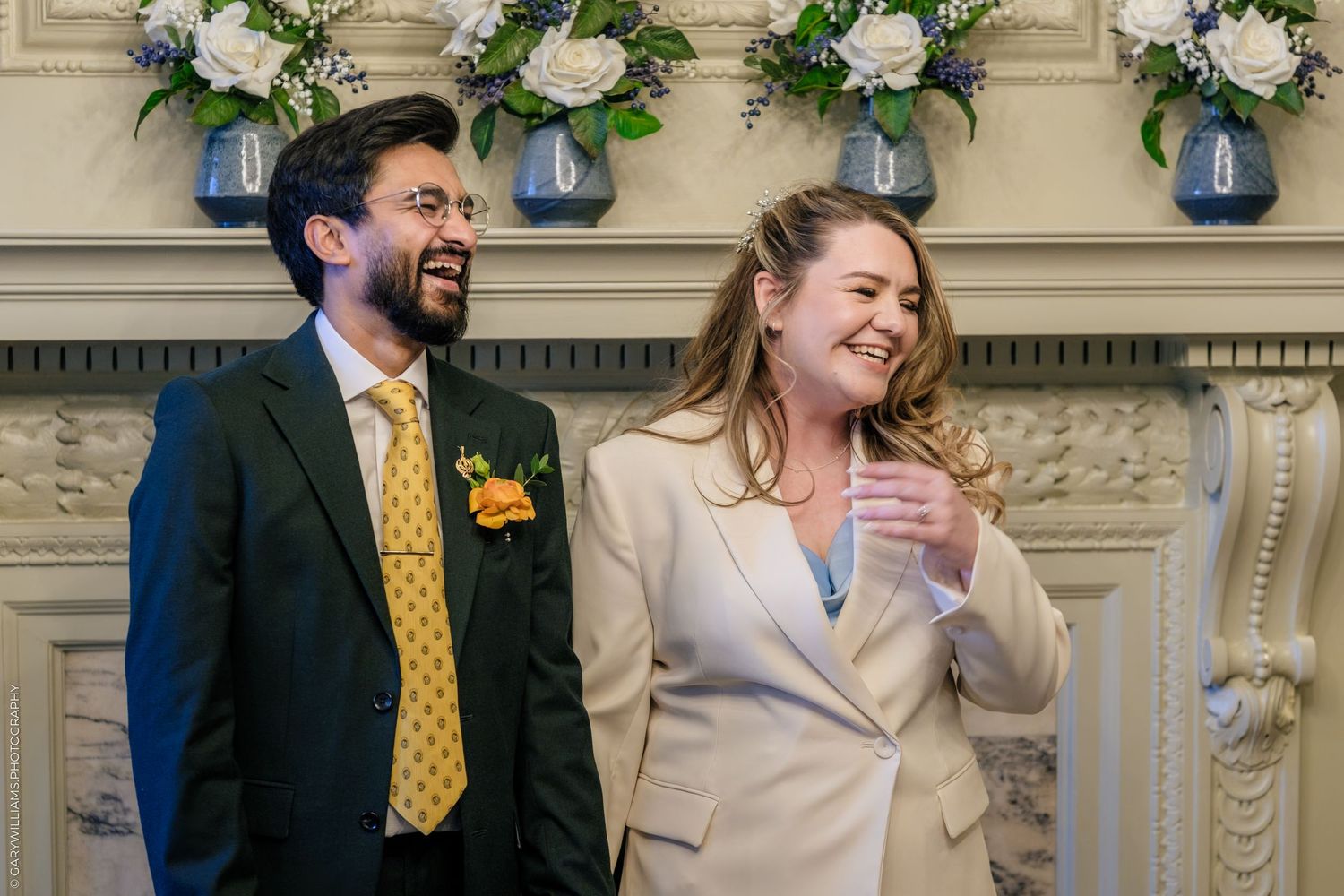 Couple laughing together at wedding ceremony, man in navy suit with yellow tie, woman in cream blazer, floral backdrop.