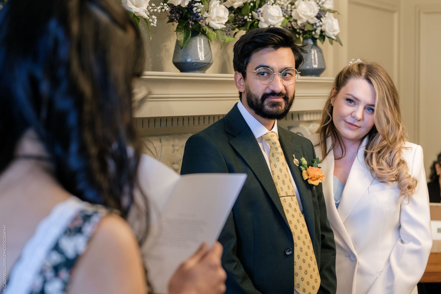 Couple exchanging wedding vows at an elegant ceremony, groom in dark suit with orange tie, bride in white jacket.