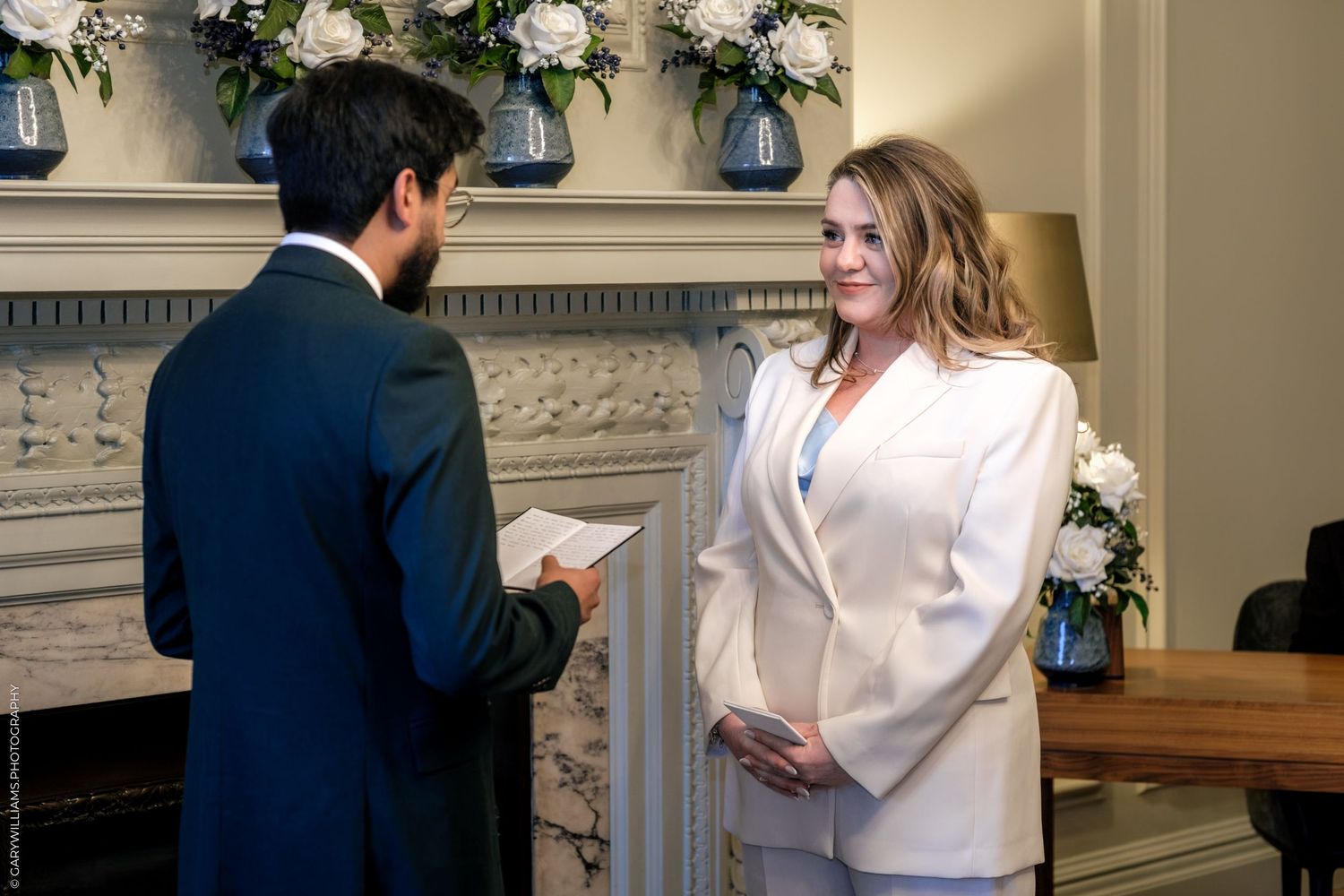 Couple exchanging wedding vows in elegant venue with white floral arrangements and classic fireplace backdrop.