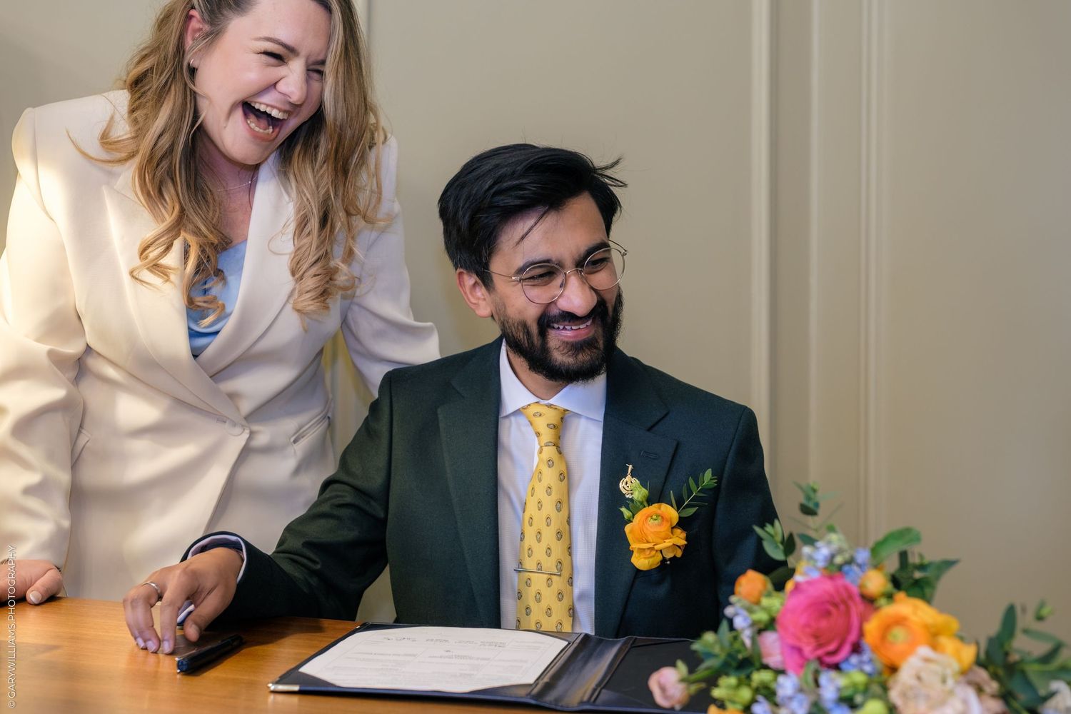 A laughing couple in formal attire sign documents at a table with colorful flowers during a wedding ceremony.