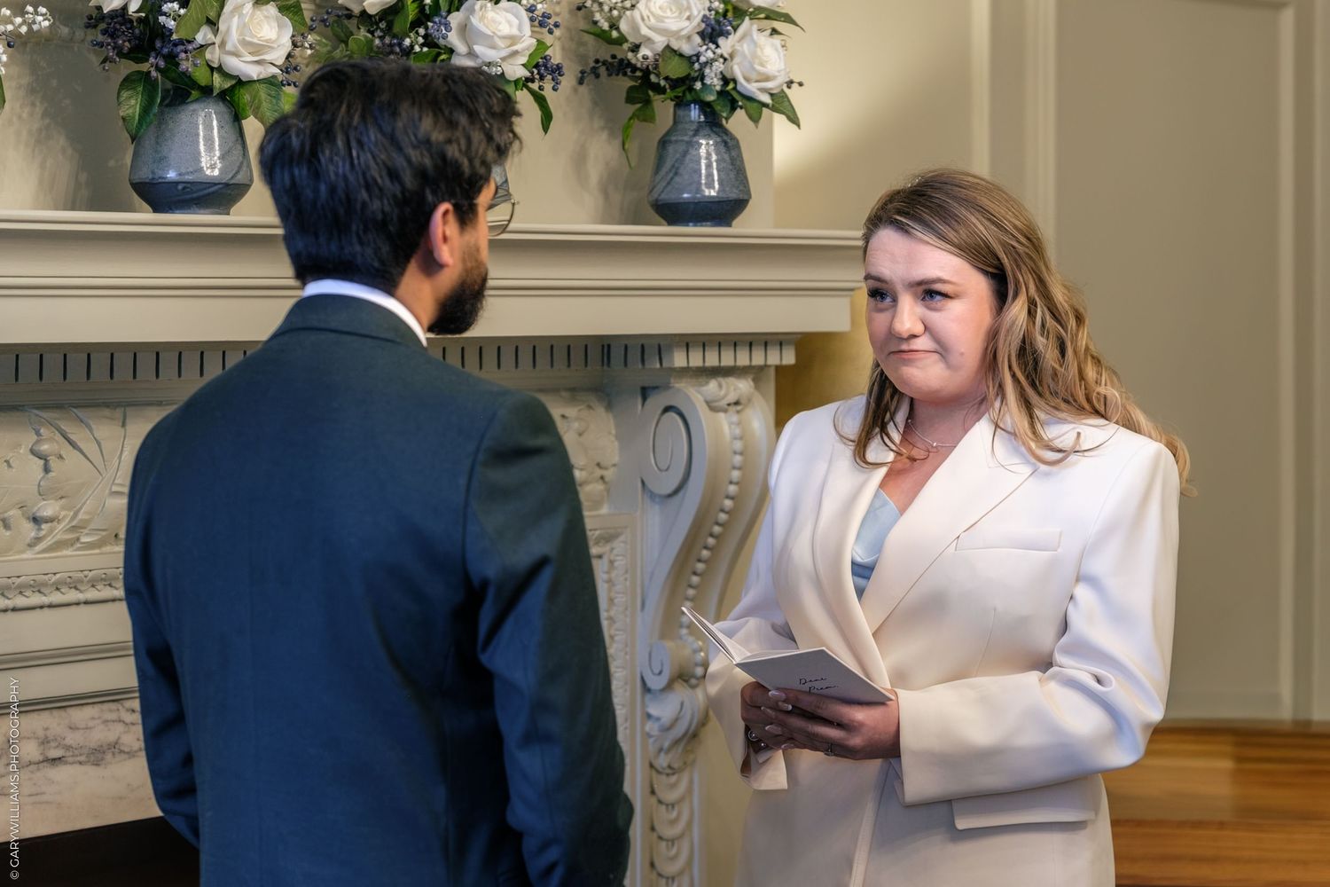 A celebrant in a white blazer reads from a book during an elegant wedding ceremony beside a decorative fireplace.
