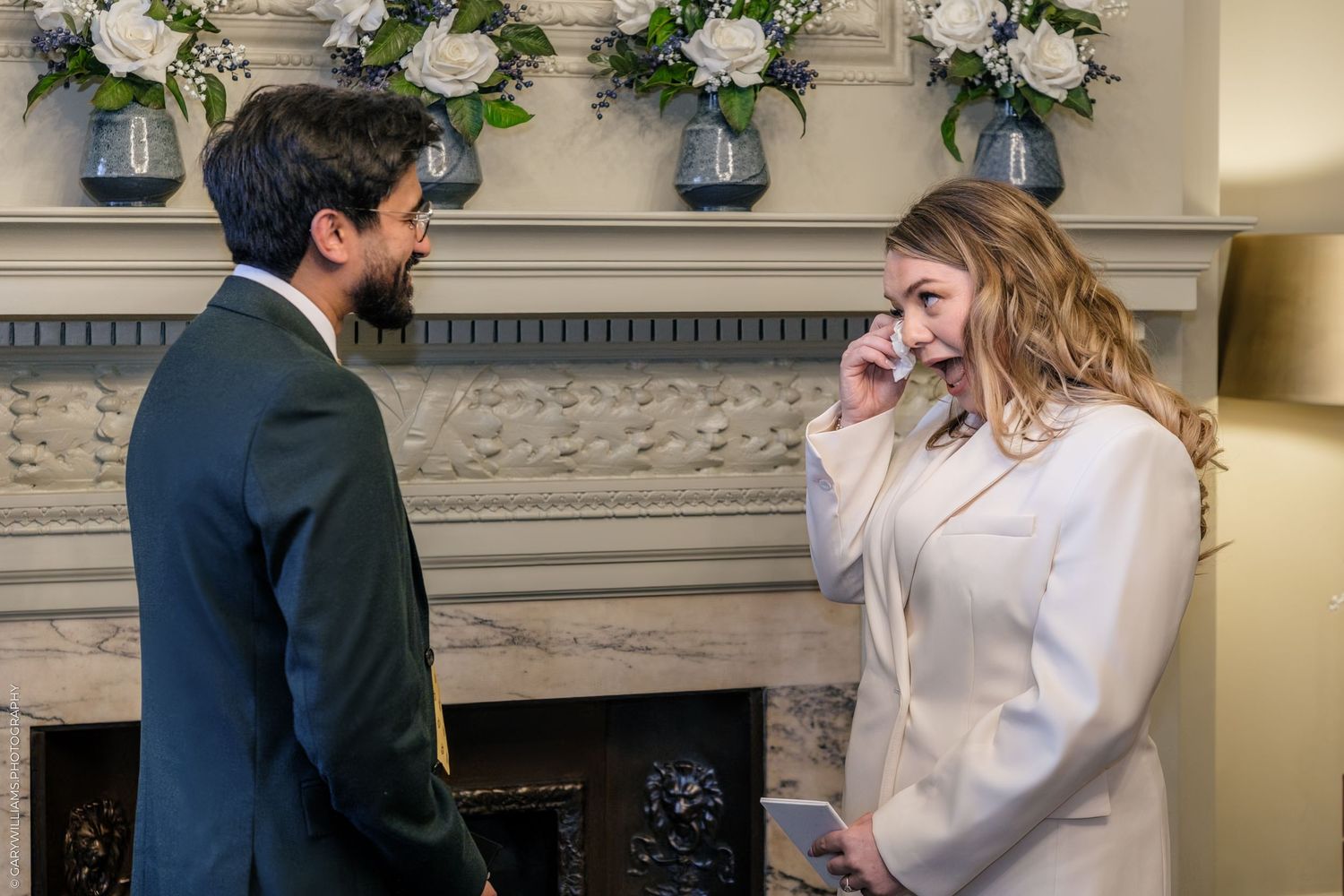 Emotional woman in white outfit wipes tears near fireplace while man in dark suit watches during wedding ceremony.