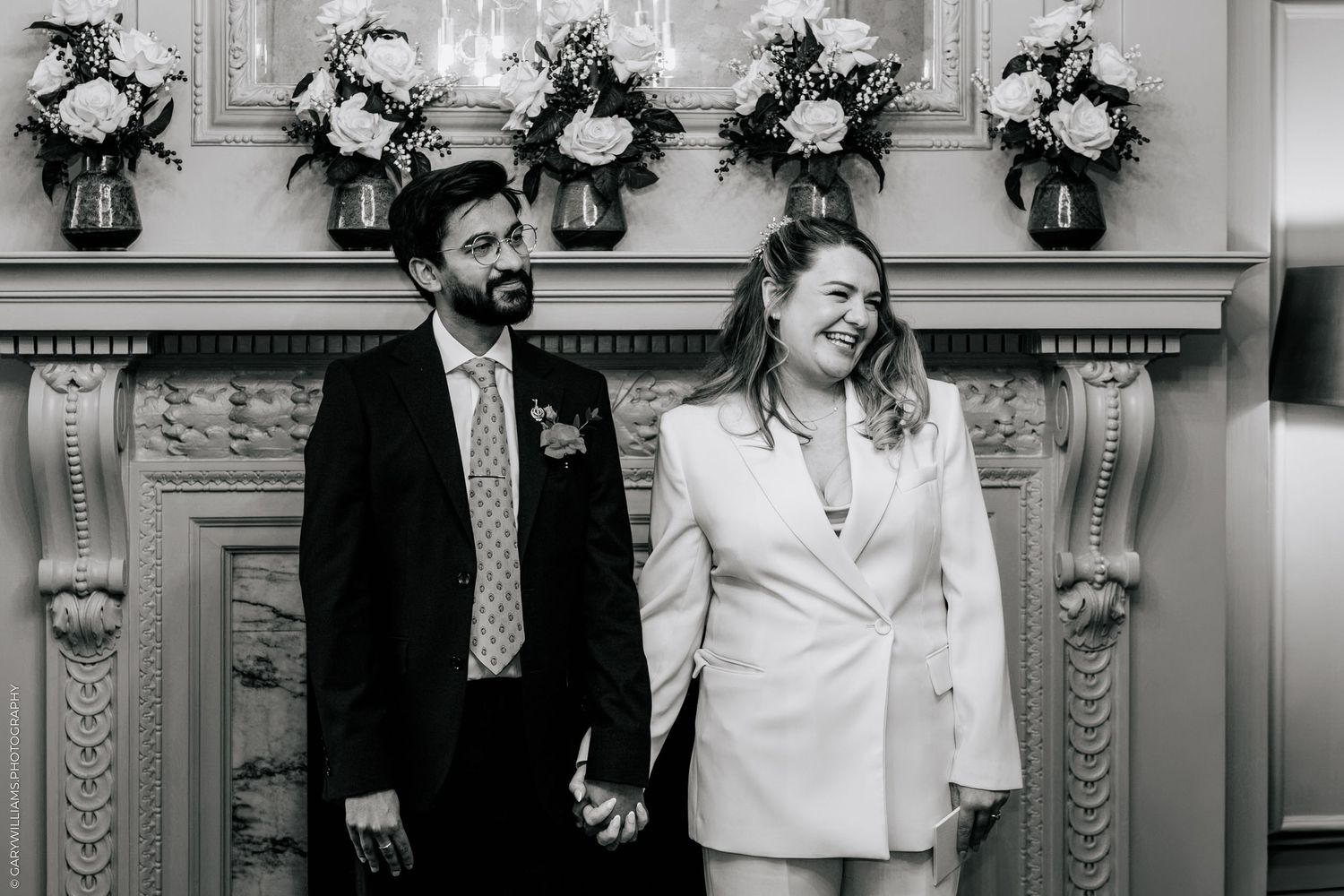 A smiling couple holds hands in front of a fireplace, groom in dark suit, bride in white blazer dress, with floral arrangements.